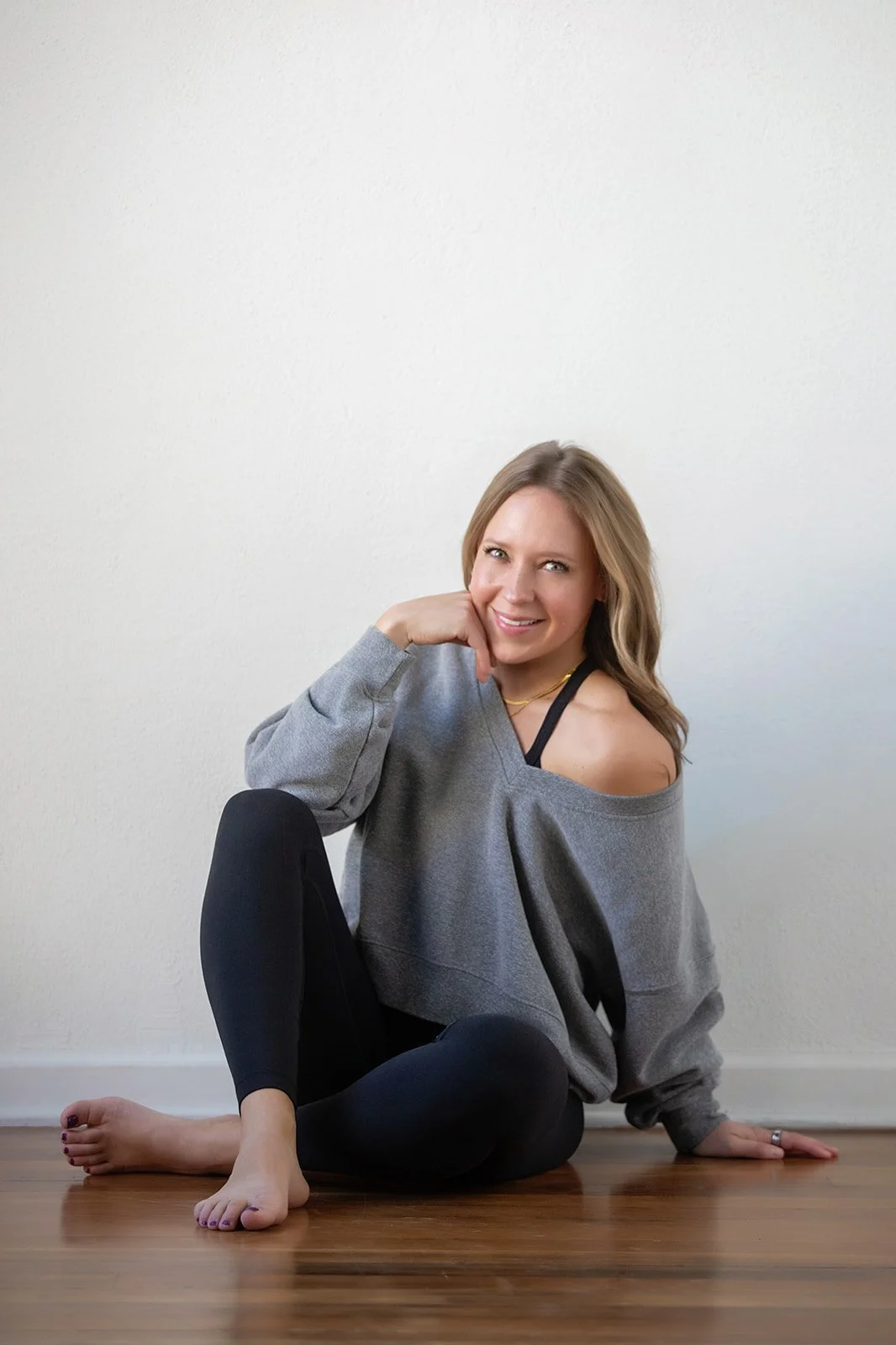 A woman with long blonde hair sitting barefoot on a hardwood floor against a plain white wall, wearing a gray off-the-shoulder sweatshirt and black leggings, smiling and resting her chin on her hand.