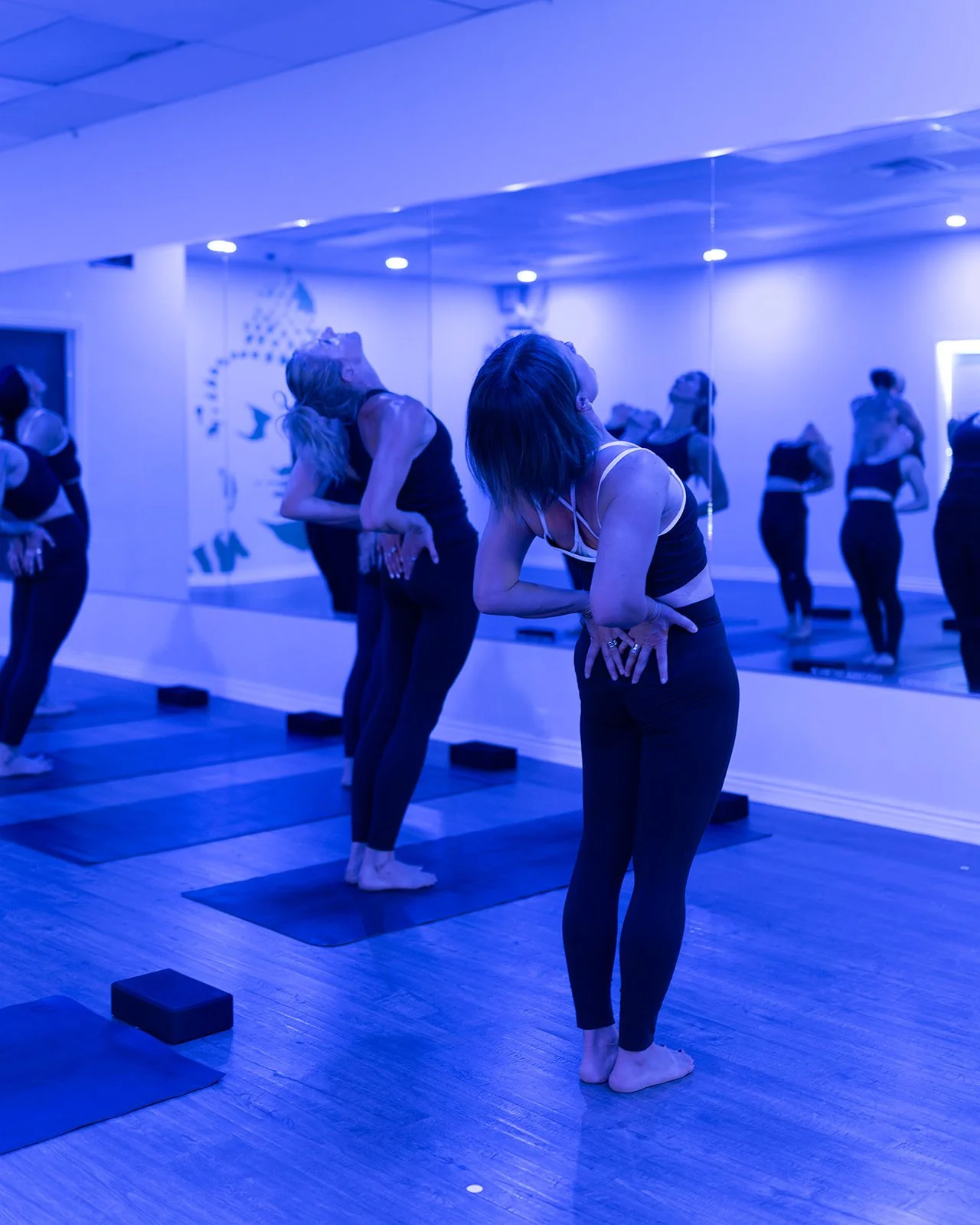 Women participating in a yoga or fitness class in a studio with mirrors, blue lighting, yoga mats, and blocks.