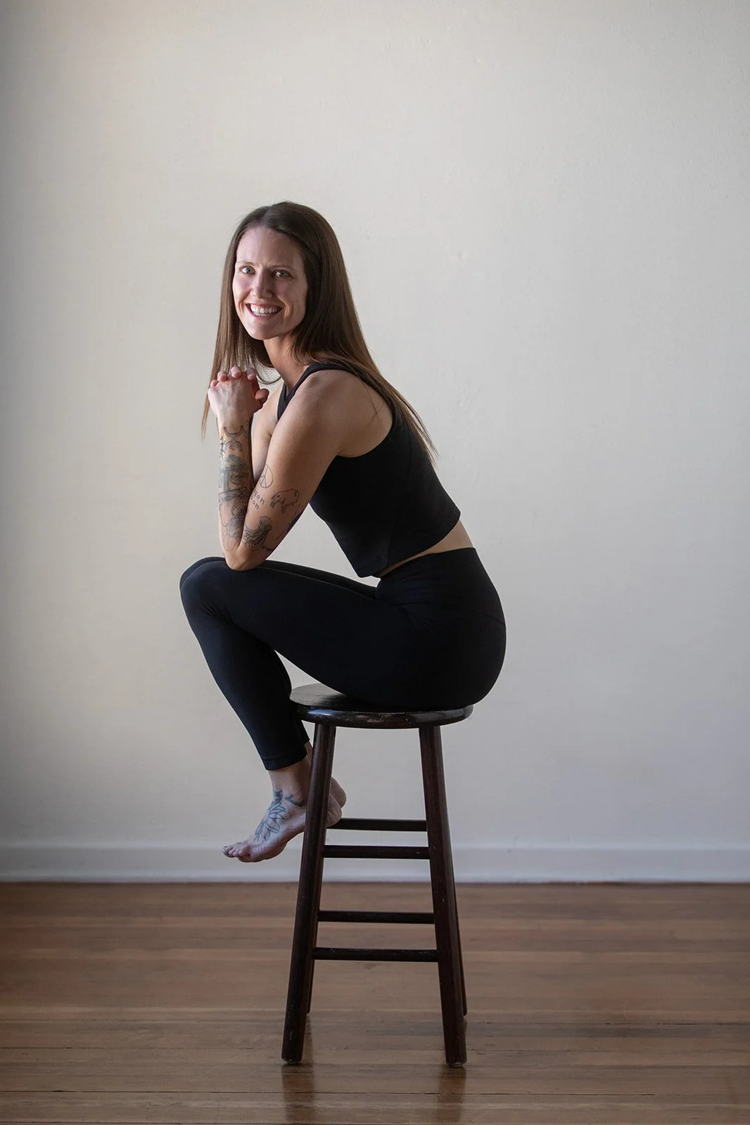 A woman with long brown hair wearing a black sleeveless top and black leggings, sitting on a wooden stool with one leg crossed over the other, smiling at the camera against a plain light-colored wall.