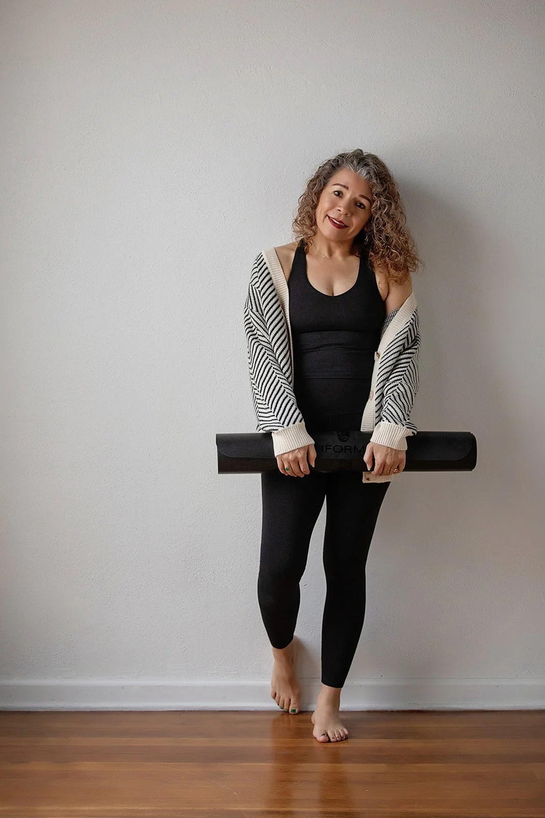 Woman with curly hair holding a black yoga mat, standing barefoot against a plain white wall.