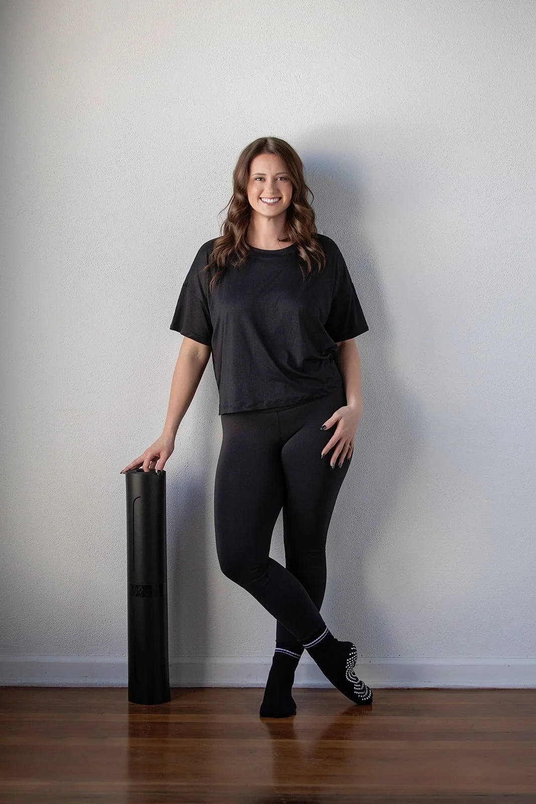 A woman in black workout clothes standing next to a black foam roller, smiling against a plain light-colored wall with a wooden floor.