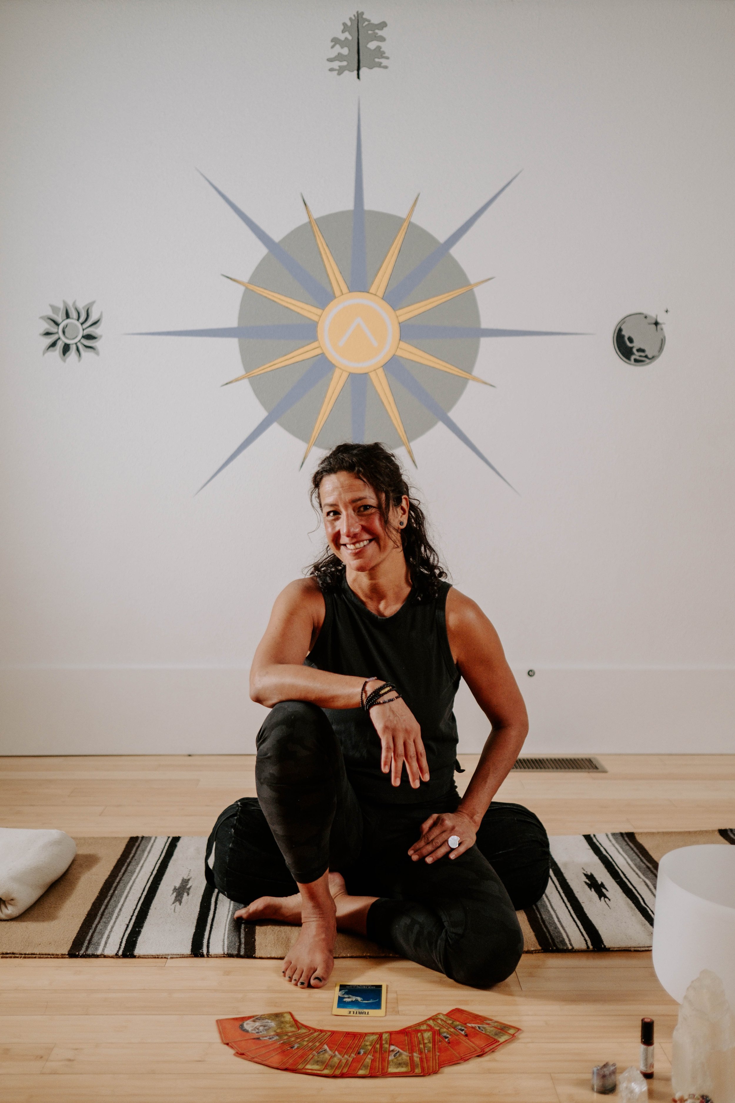 A person sitting cross-legged on a mat in front of a wall with a large sunburst design. There are tarot cards laid out on the floor and various small objects next to them. The person is smiling and wearing a sleeveless black top and dark leggings.