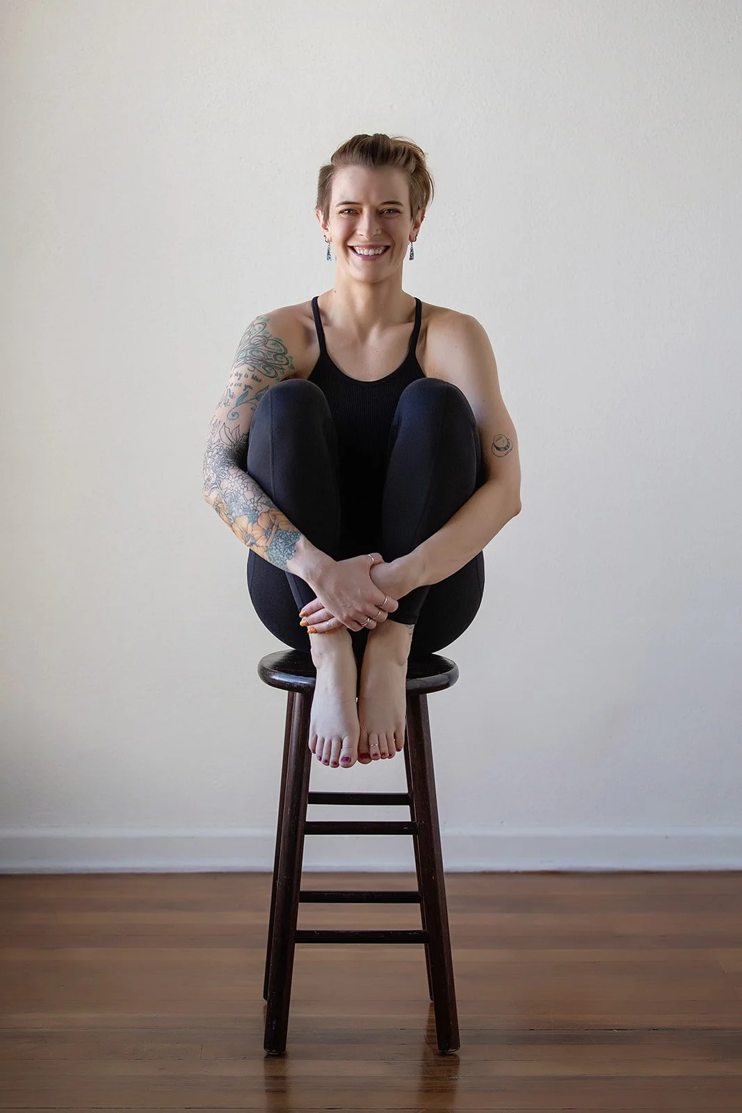 A woman with tattoos on her arms is smiling while sitting on a stool with her knees drawn to her chest and her arms wrapped around her shins, against a plain white wall.