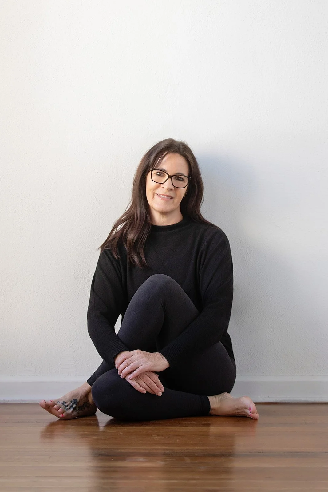 Woman sitting on wooden floor in front of a plain white wall, wearing black clothing and glasses.