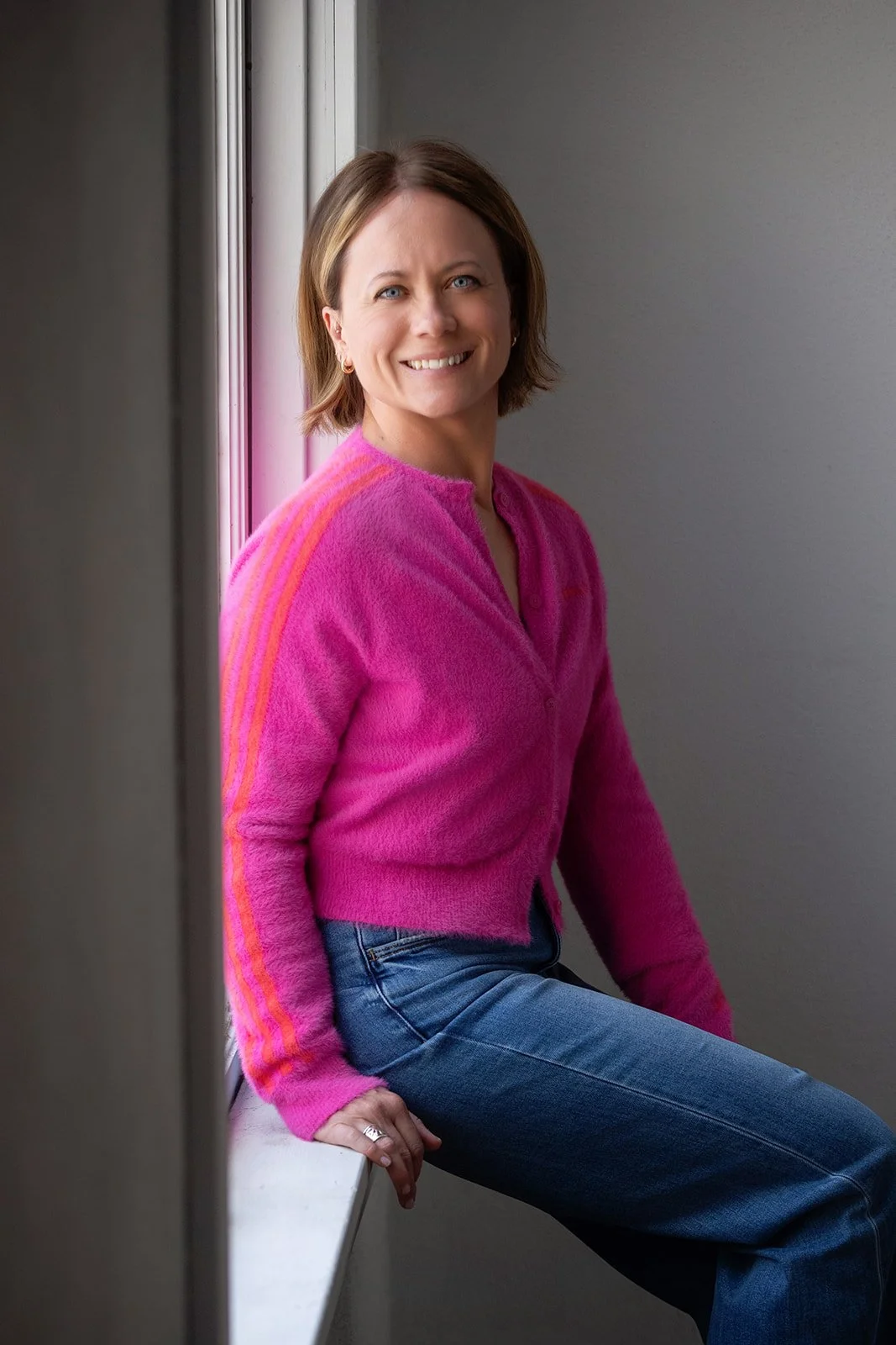 A woman with short brown hair wearing a pink sweater and blue jeans, sitting by a window and smiling.