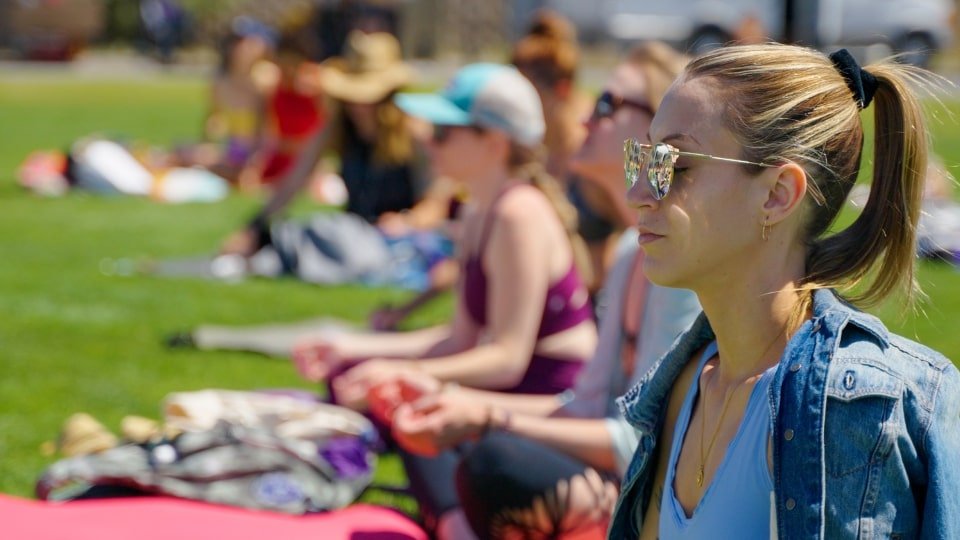 Group of people sitting on grass, meditating outdoors, wearing casual clothing and sunglasses.