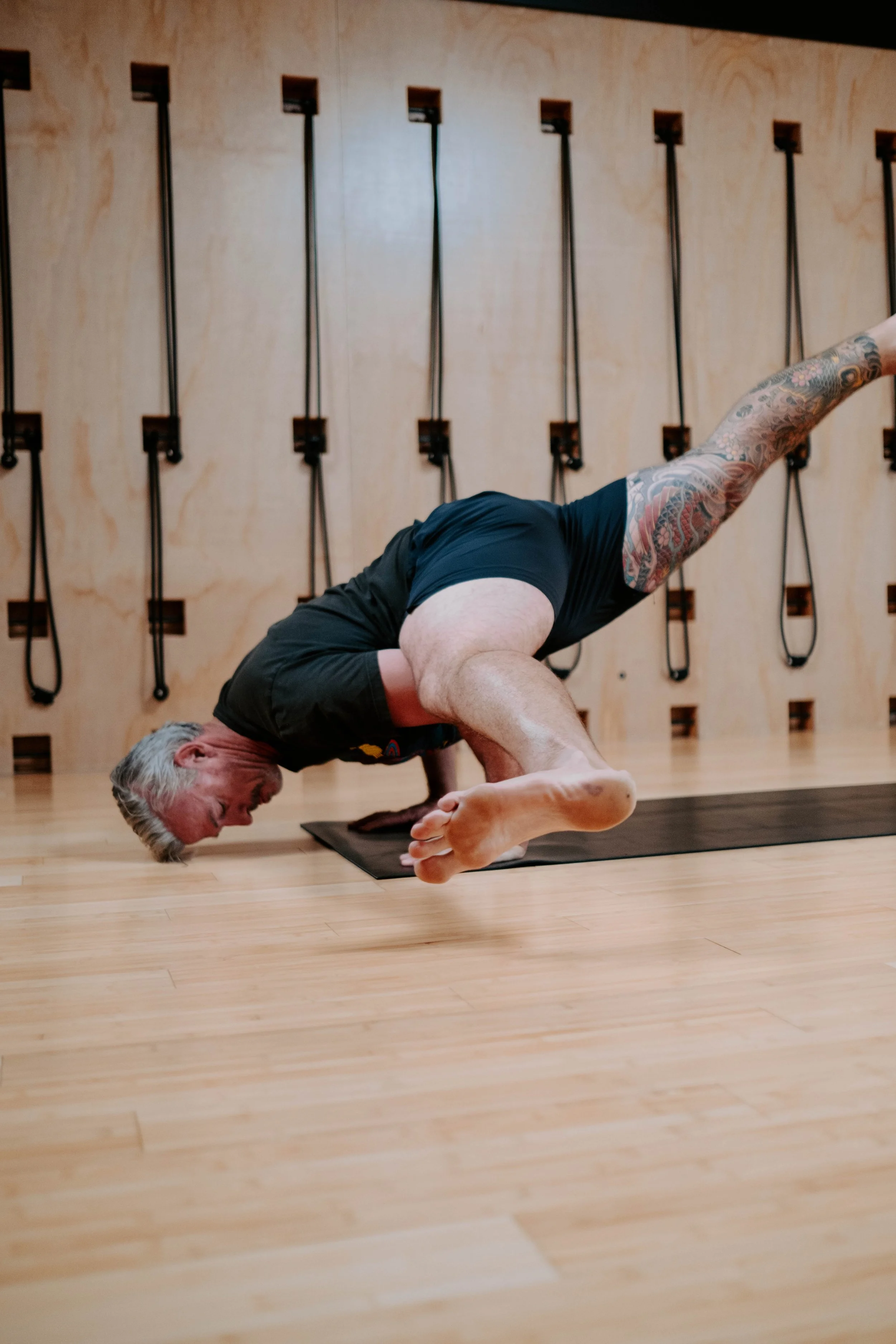 Man performing advanced yoga pose on mat indoors with tattooed arm and leg, wooden wall and straps in background.