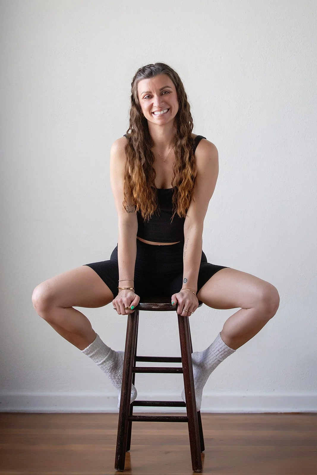 A young woman with long, wavy brown hair, sitting on a wooden stool with her legs spread apart, smiling and looking at the camera, wearing a black tank top, black shorts, white socks, and minimal jewelry, against a plain white wall.
