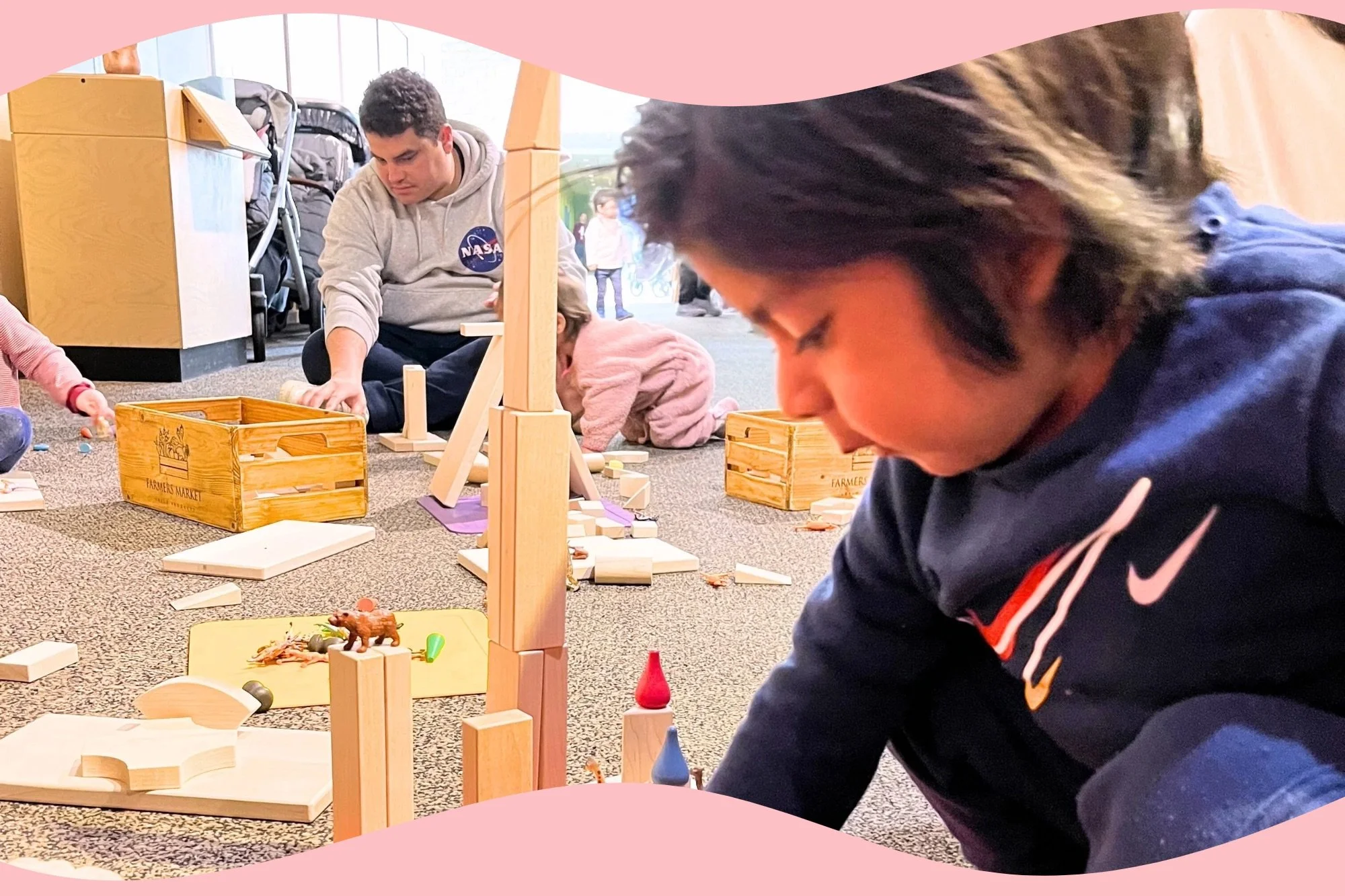 Children and an adult playing with wooden blocks and toys on the floor in an indoor play area. The children are focused on their activities, with one child close to the camera and an adult wearing a NASA hoodie in the background.