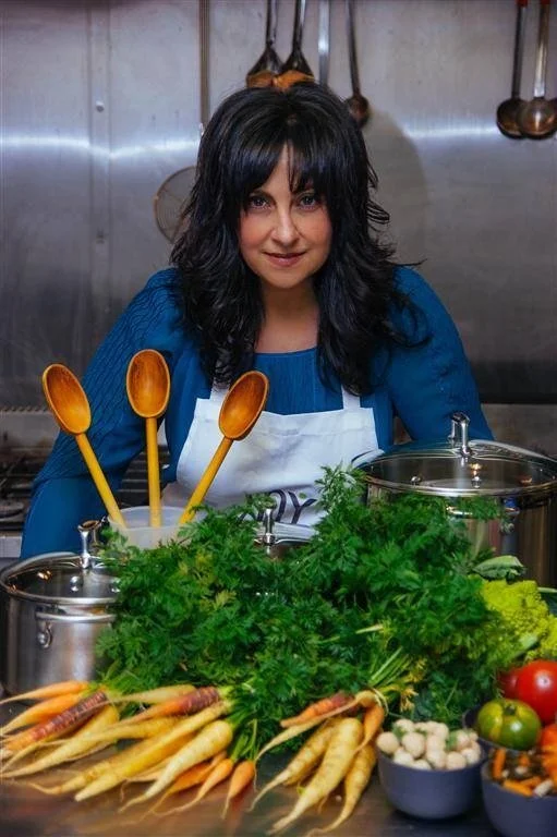 Woman with dark hair wearing a blue top and white apron standing behind a kitchen counter with fresh vegetables and cooking utensils, smiling at the camera.
