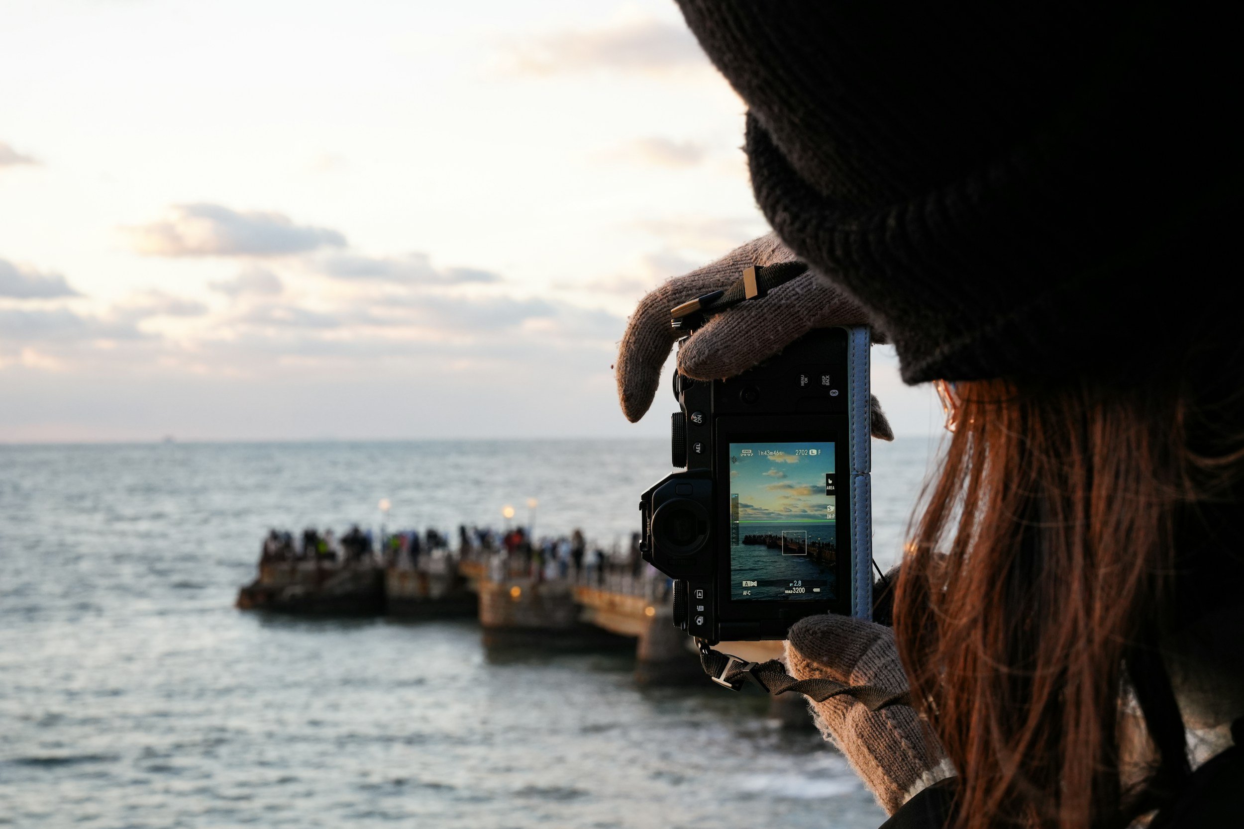 A person taking a photo of a sunset over the ocean with a digital camera, on a pier with a group of people in the background.