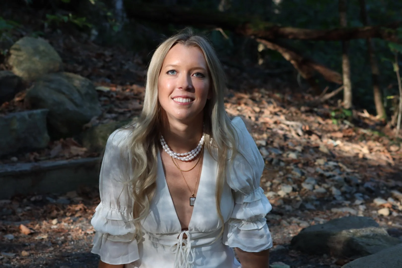A woman with long blonde hair wearing a white blouse with puffy sleeves, layered pearl necklaces, and a gold pendant necklace stands outdoors in a wooded area with rocks and fallen leaves.