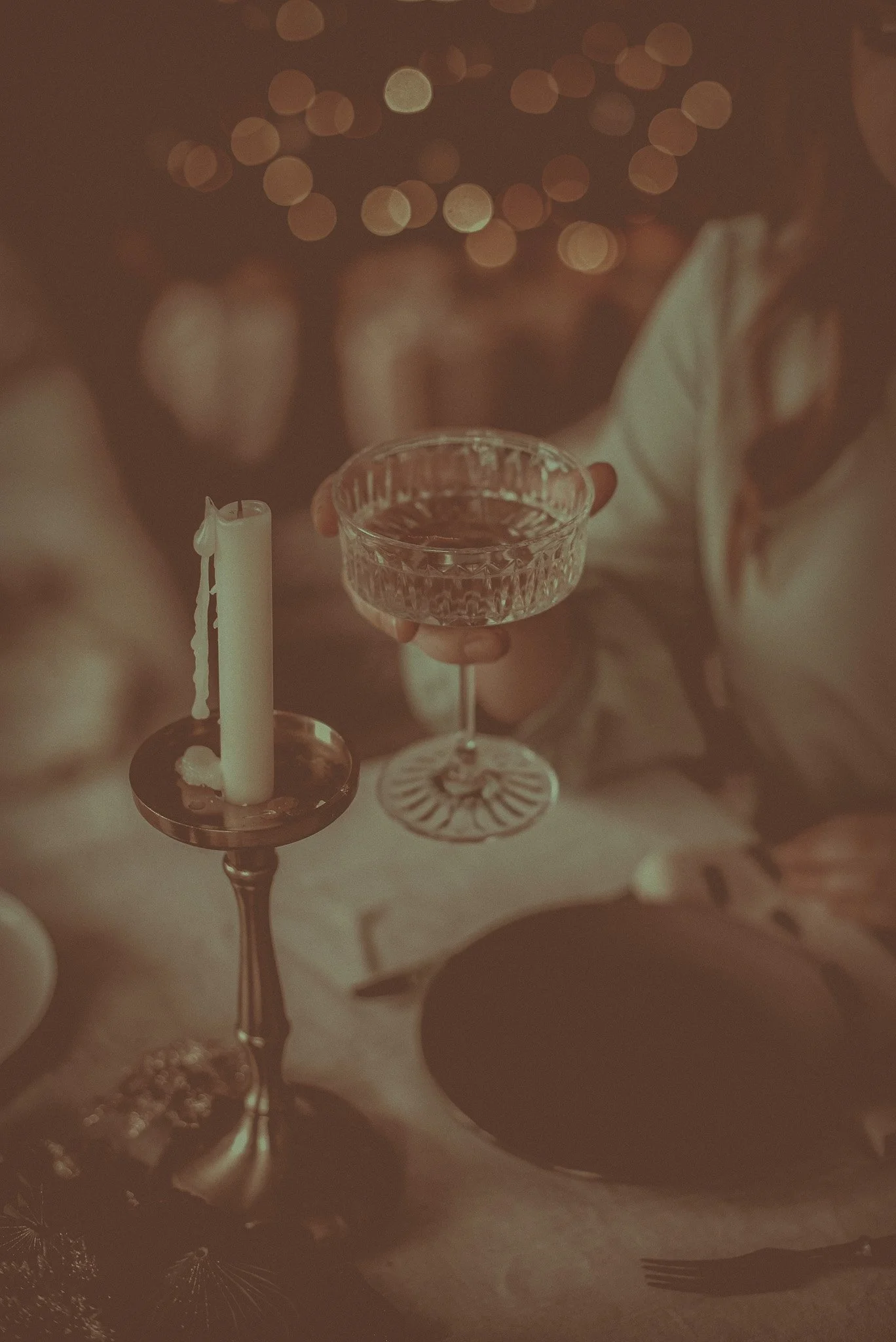 Person holding a vintage glass of drink at a dimly lit dinner table with a lit candle in a candle holder.
