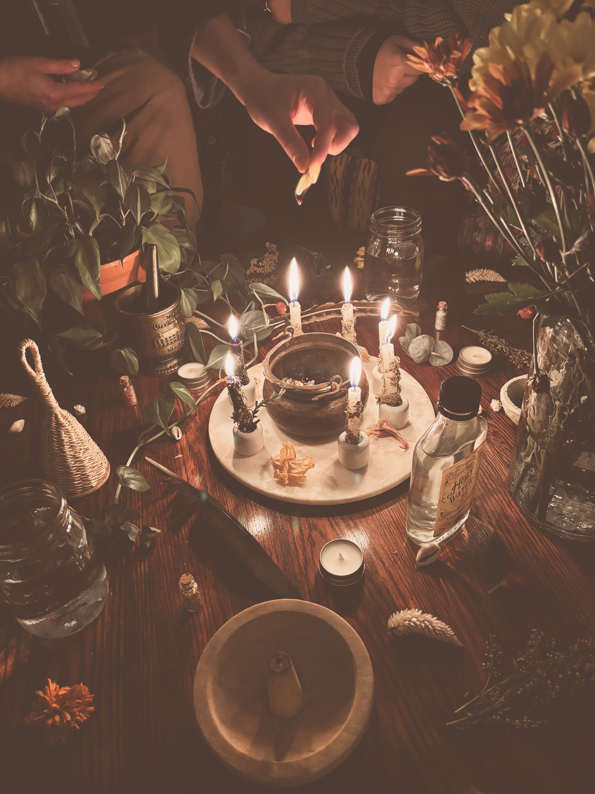 An altar with plants, feathers, bells, holy water, incense, flowers, mortar and pestle, cauldron and candles surrounded by a group of witches performing a ritual together.
