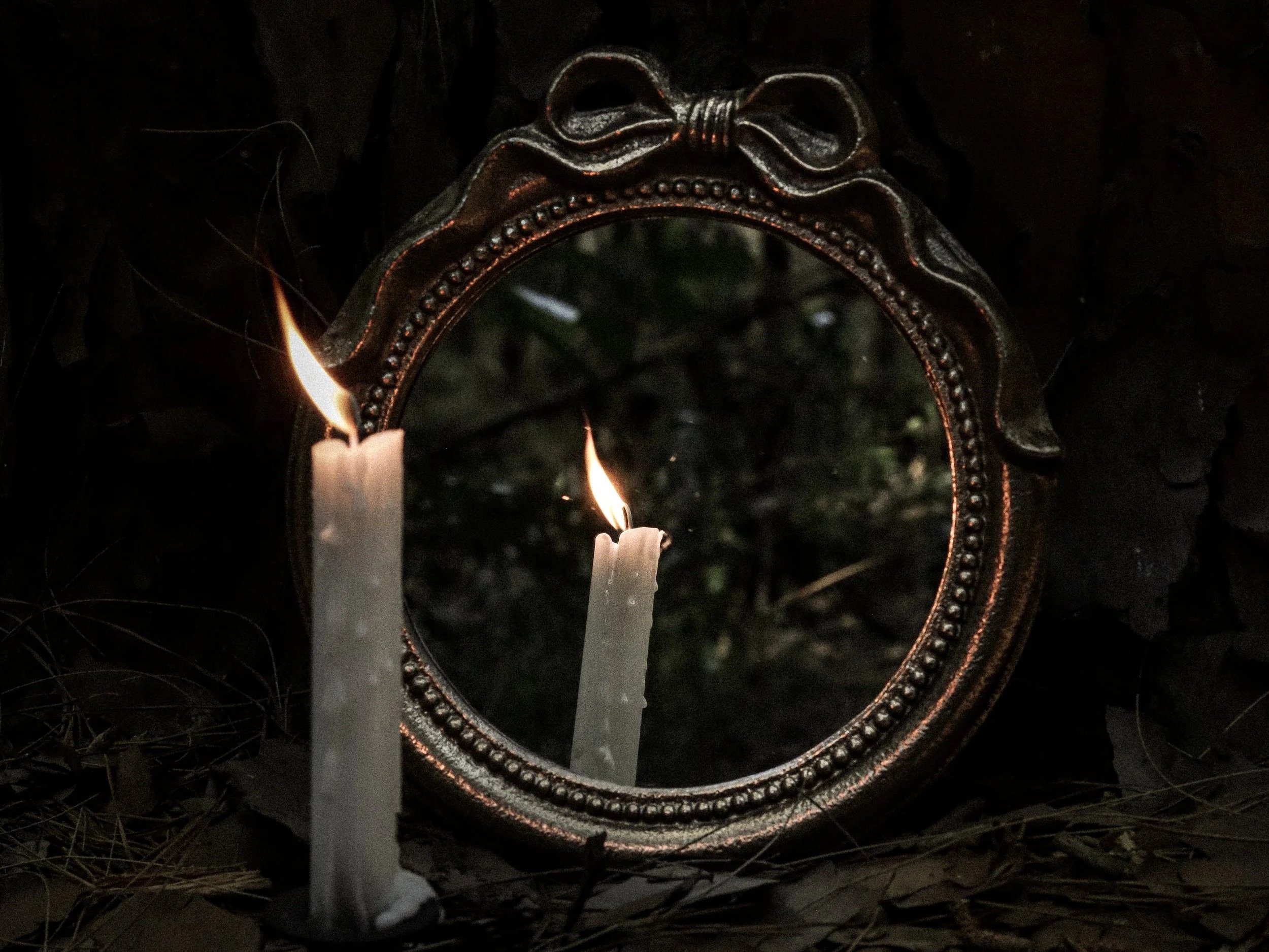 A white taper candle burns set on the ground outdoors among decaying leaves with a vintage-looking round mirror with a gold frame with beading and a ribbon reflecting the candle back.