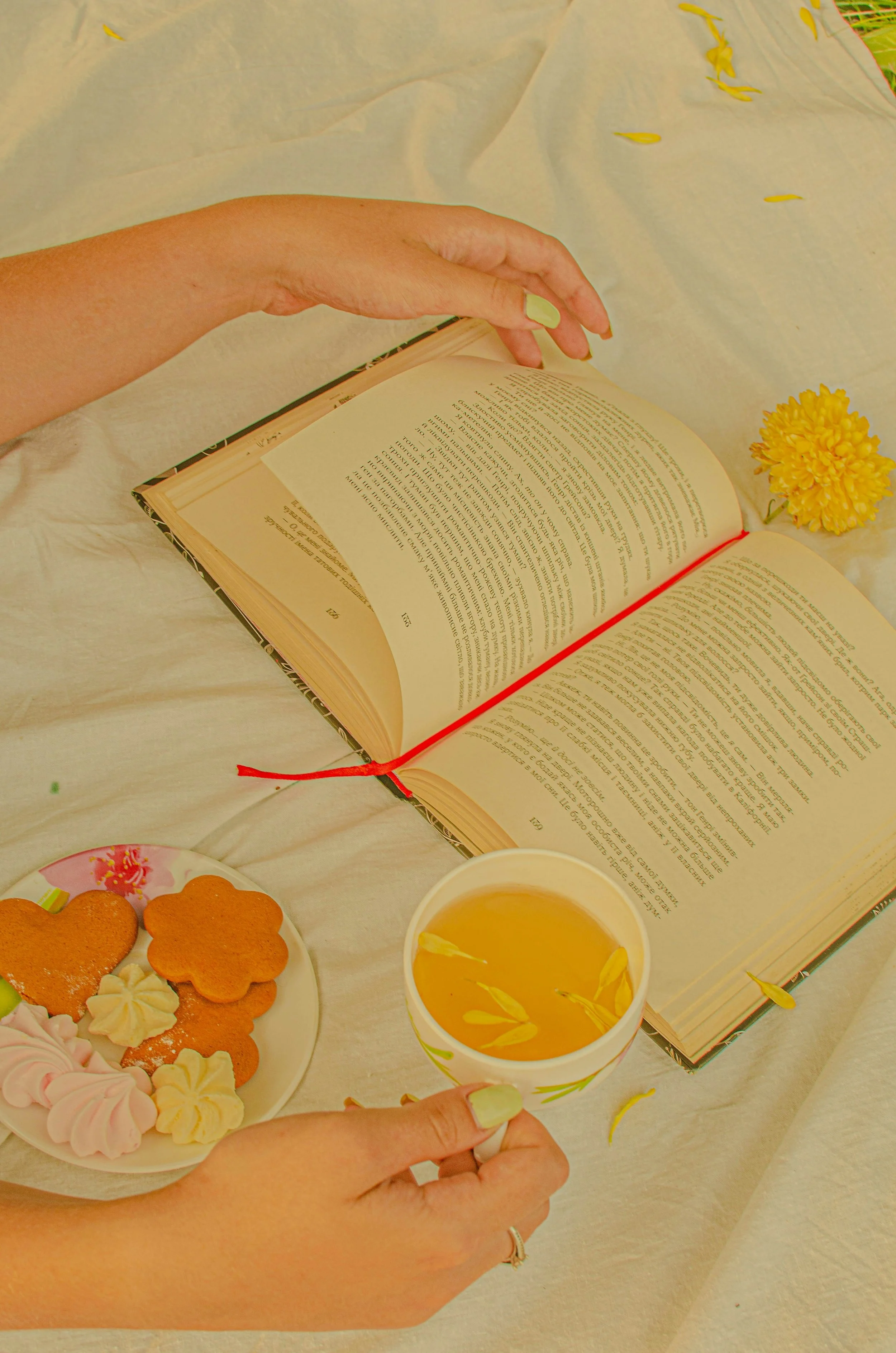A golden, vintage light over a photo of a woman's hand with bright celery green nail polish turning the page of a book. There aer orange, pink, and yellow cookies with a cup of tea and some yellow flowers and petals.