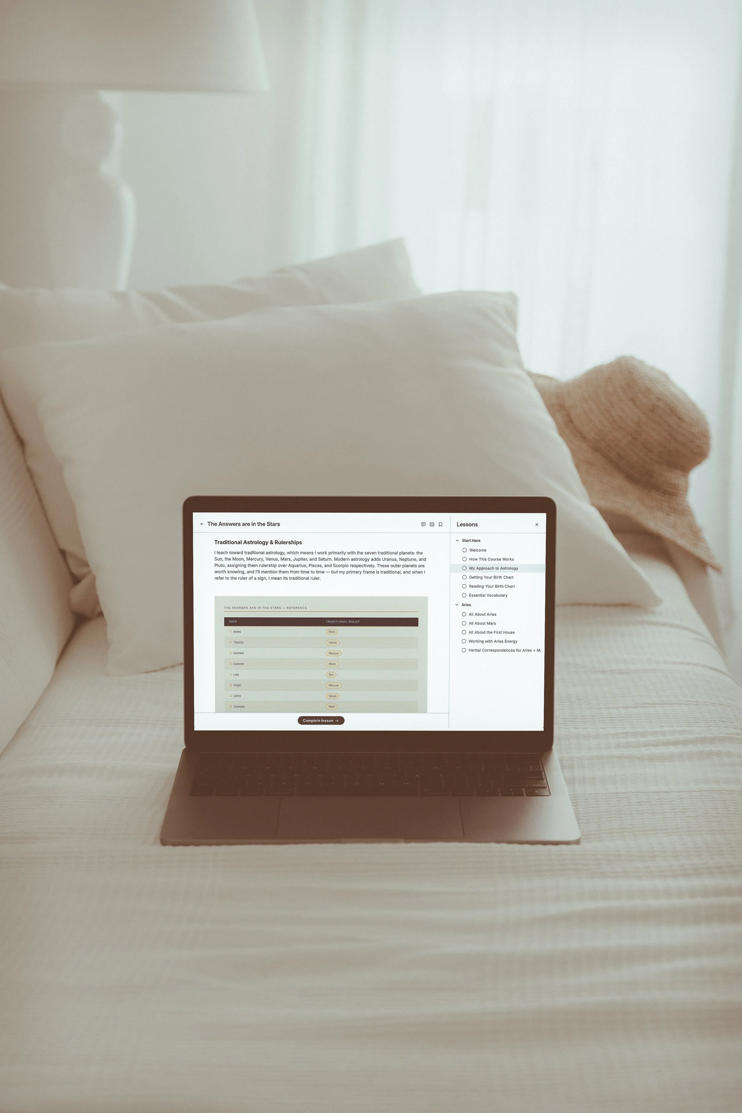 A laptop sits on a daybed with an astrology course on the screen. There are pillows and a garden straw hat on the bed behind the laptop.