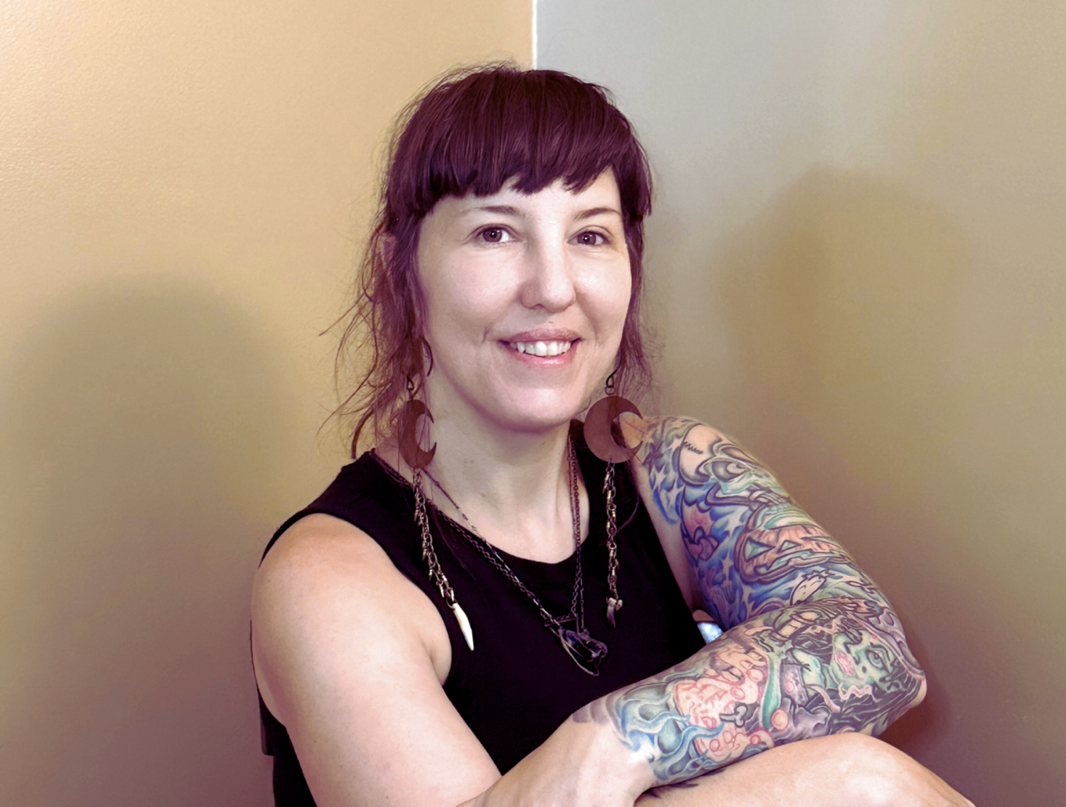 A woman with short reddish-purple hair and large moon-shaped earrings, smiling and sitting with her arms crossed, displaying colorful tattoos on her arms, wearing a black sleeveless top, against a plain beige wall.