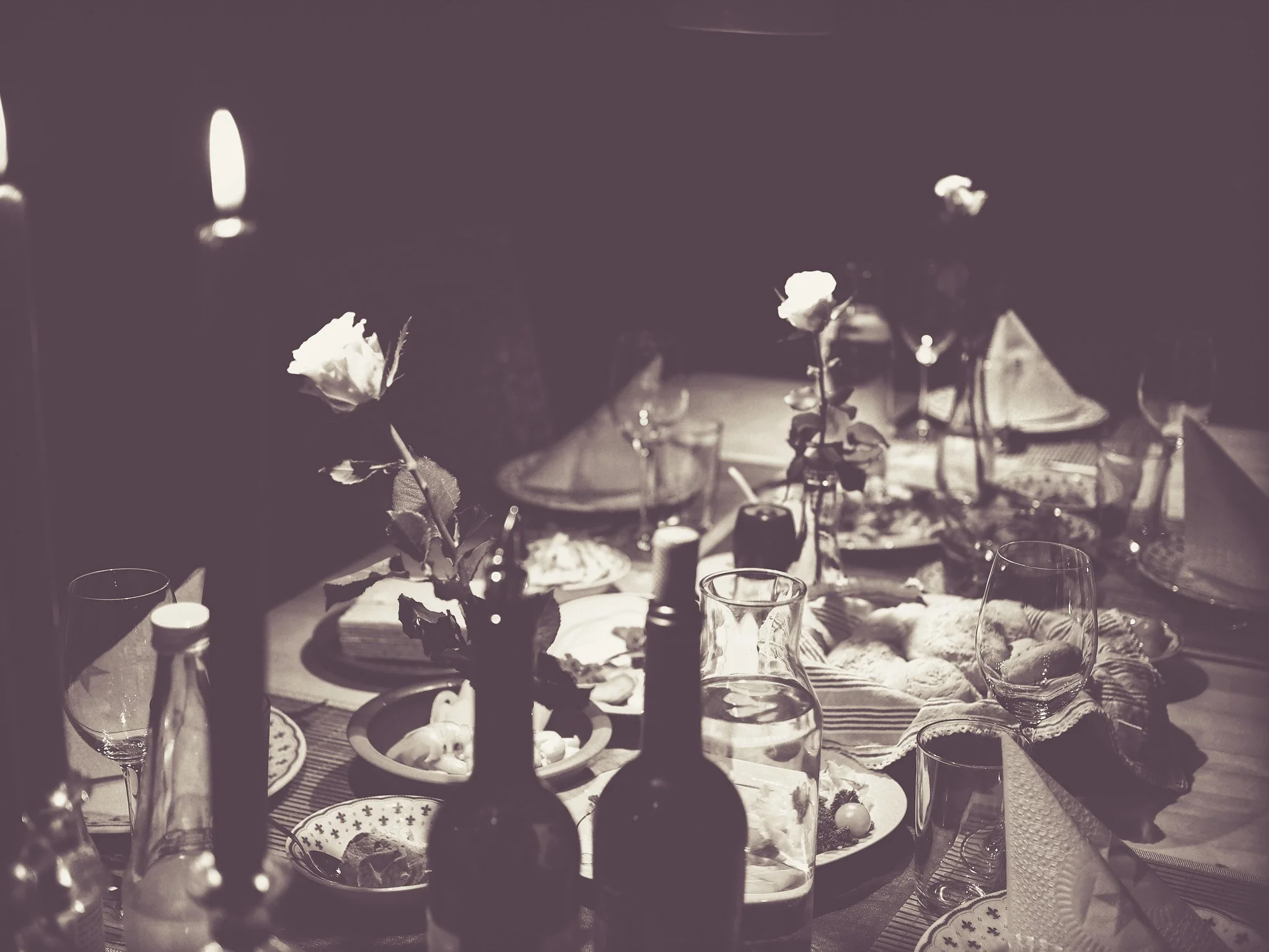 A table set for a dinner party with plates, glasses, bottles, flowers, and candles, with a dark background.