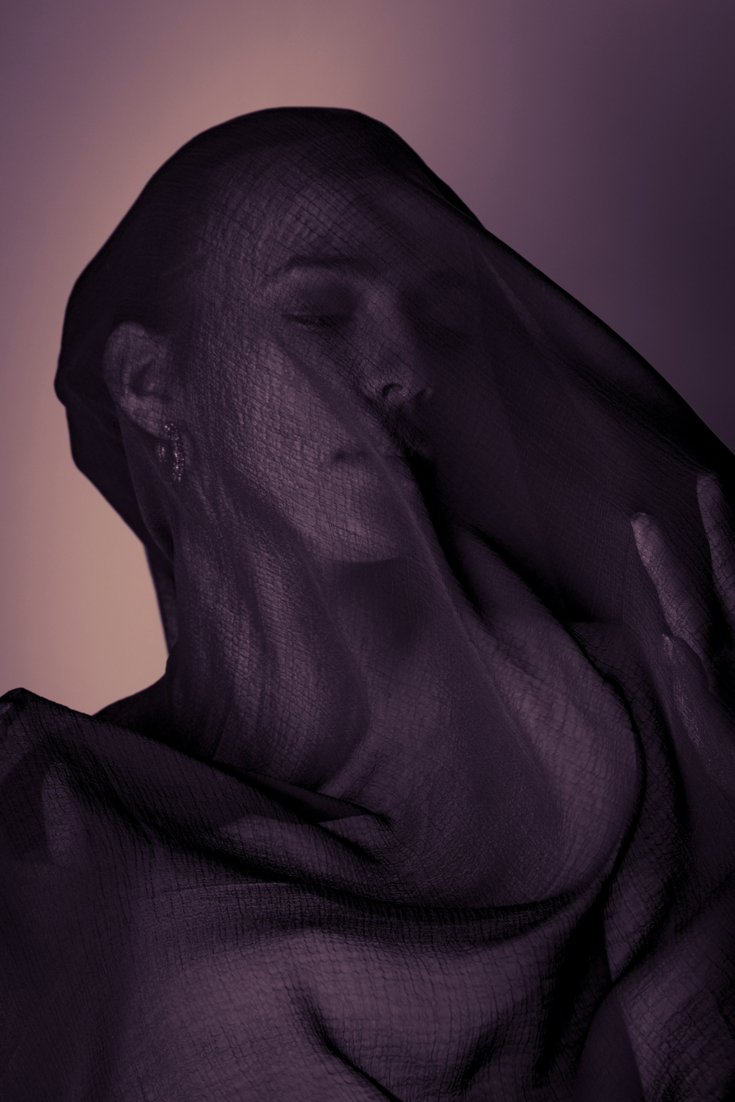 A photo of a woman with a dark veil draped over her head and covering her face with a deep purple hue cast on the entire photo and some warm orangish notes in the background form the light.