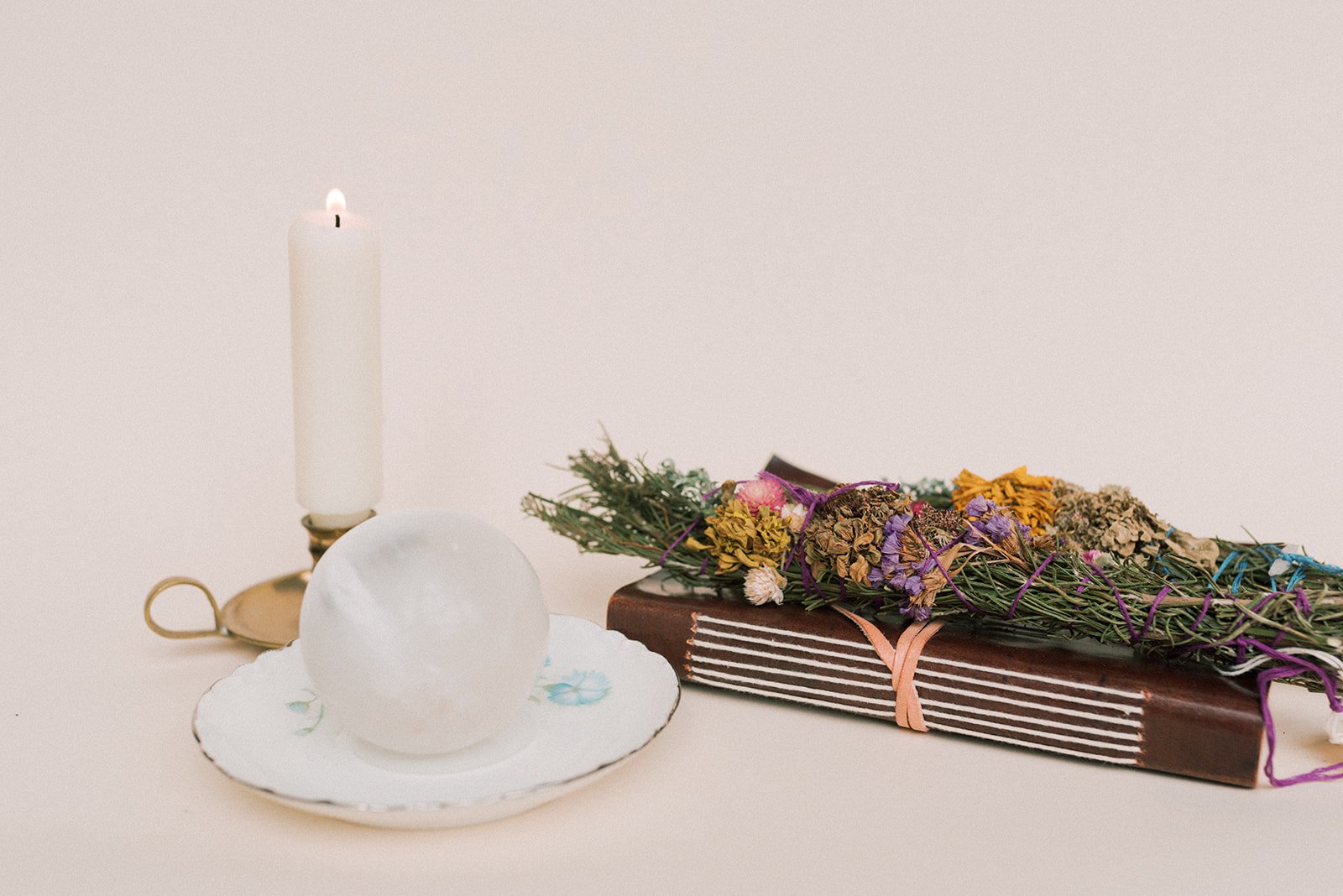 An altar-type scene with a selenite sphere resting on a vintage tea saucer with a candle in a brass candlestick holder, a leather-bound grimoire, and herbal burn wands with dried flowers
