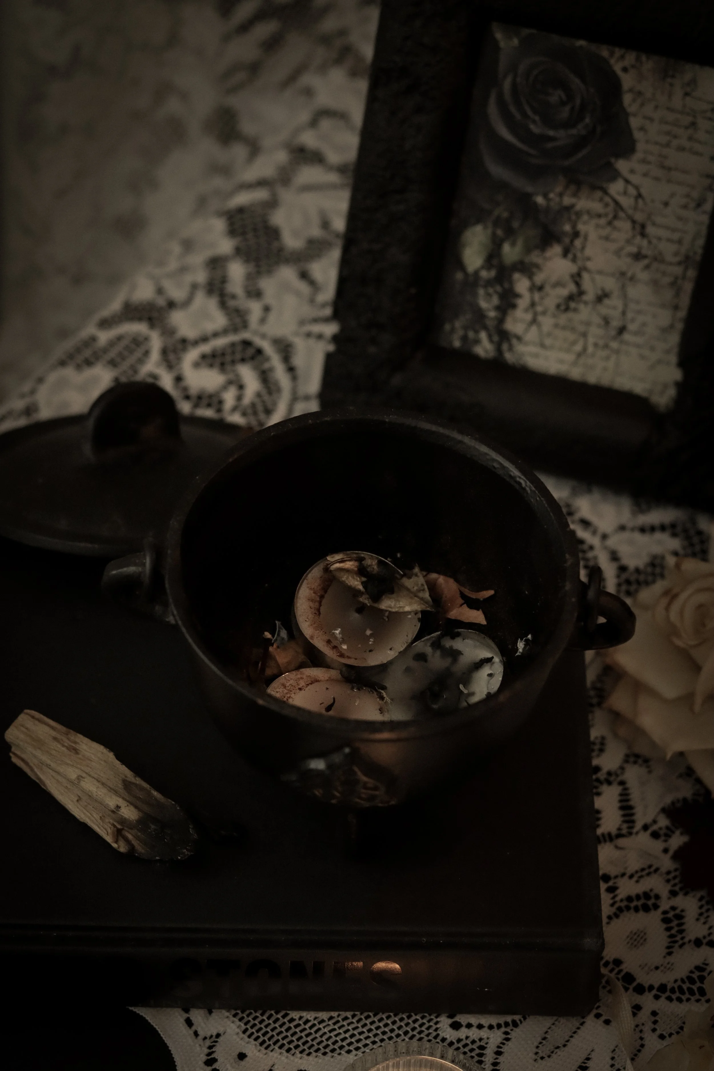 A cauldron sits on a lace tablecloth in this dark and moody photo. Tea light candles and burnt herbs rest in the cauldron with an image in a black frame just behind it of some handwriting and a black rose collaged on top.