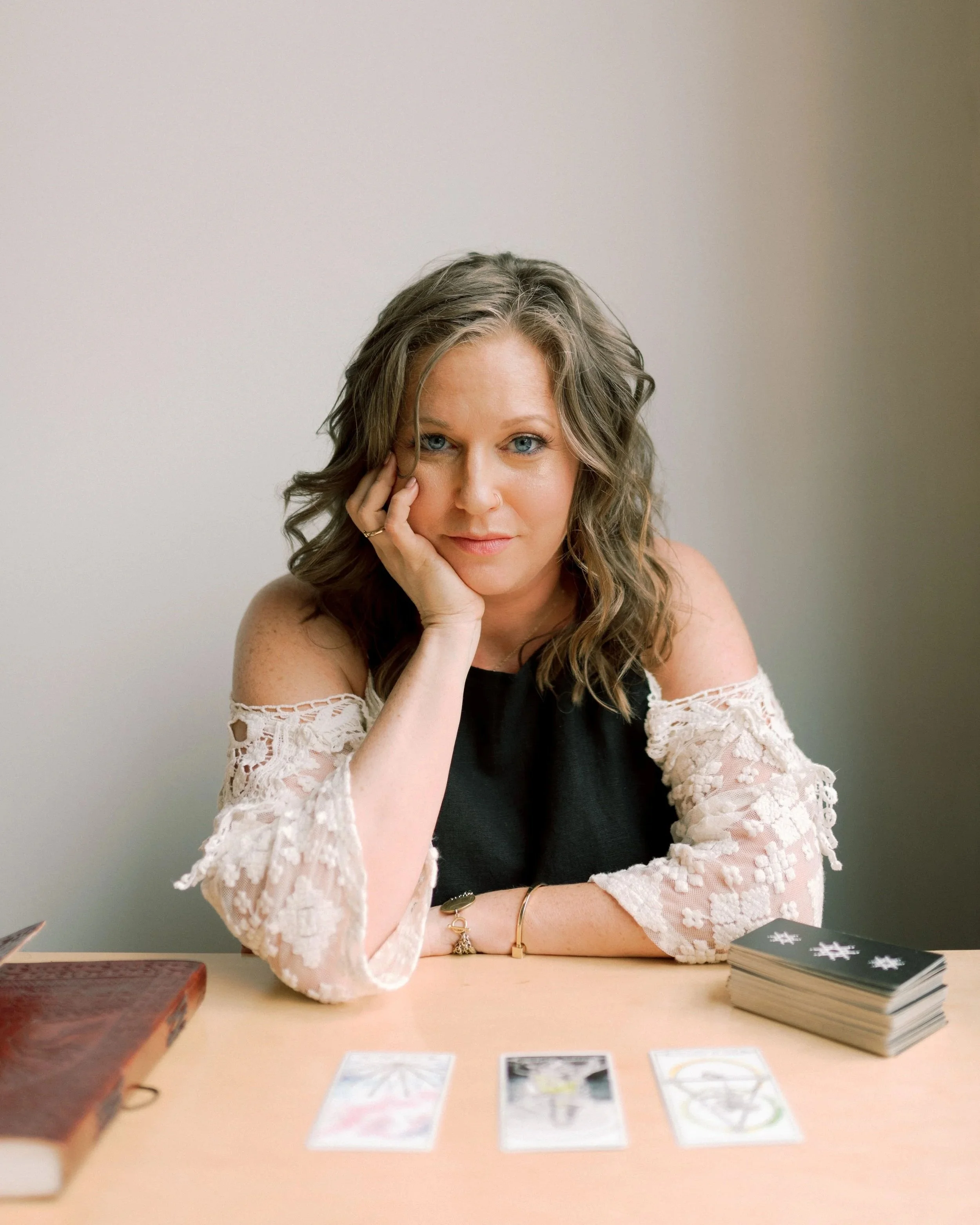 Woman with wavy brown hair sitting at a table with tarot cards and a deck, resting her chin on her hand, looking at the camera.
