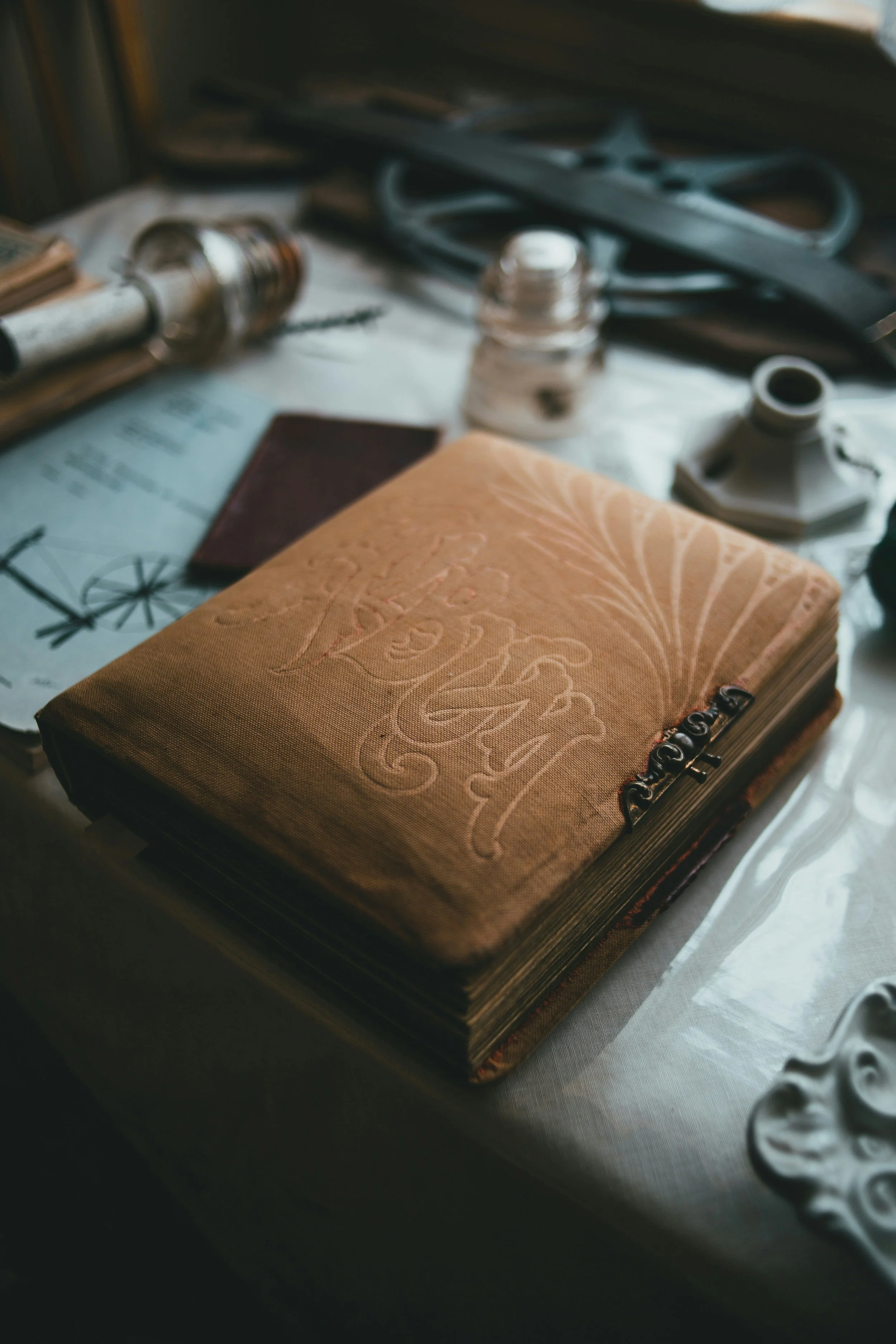 A beautiful tan linen covered book that looks old and well worn with some decorative scrolling work debossed on the fabric and an intricate metal closure in the foreground with a soft focus on the full desk with different tools and sketches.