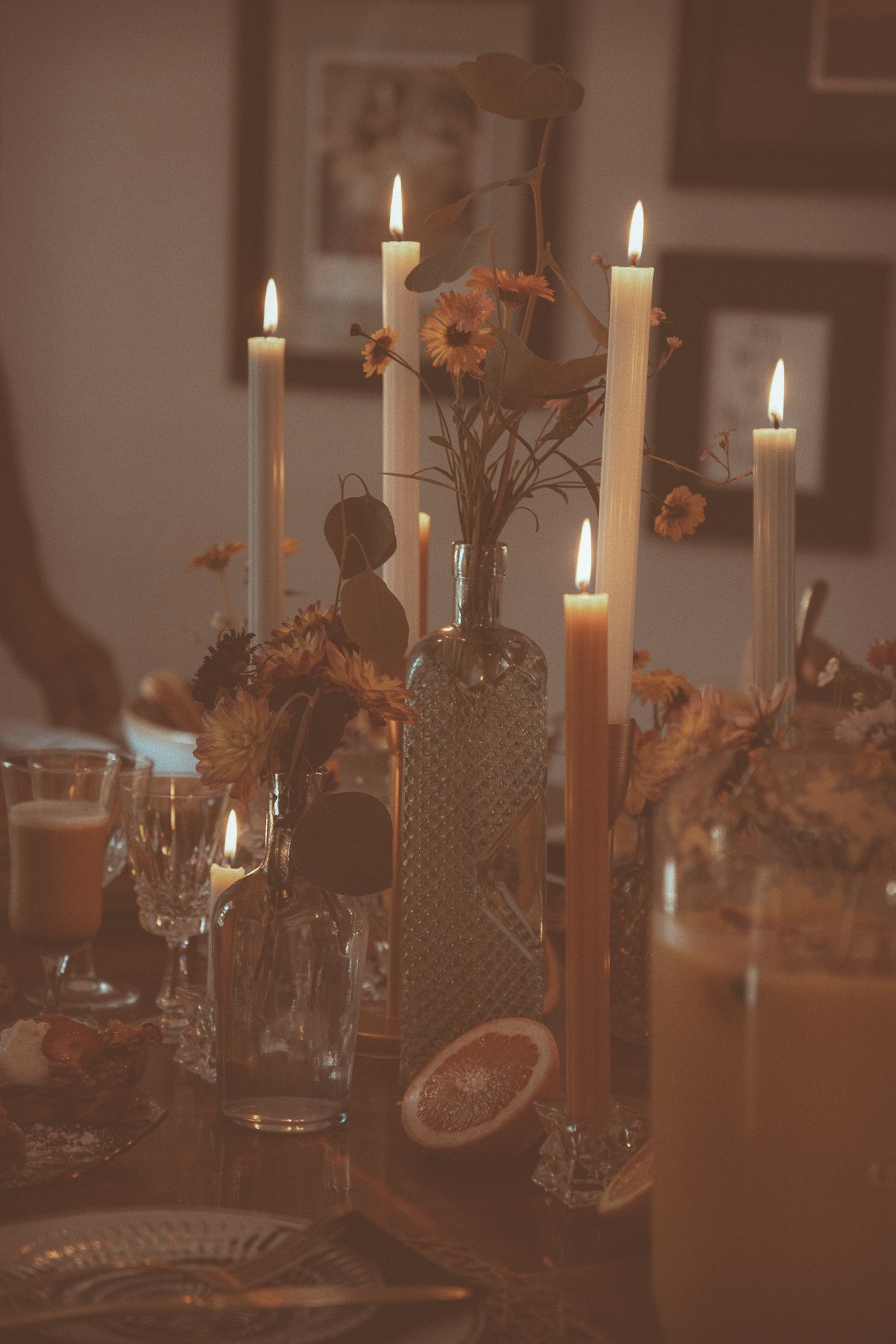 A dimly lit dinner table decorated with vases of flowers and tall candles, with a sliced grapefruit in the foreground and framed pictures on the wall in the background.