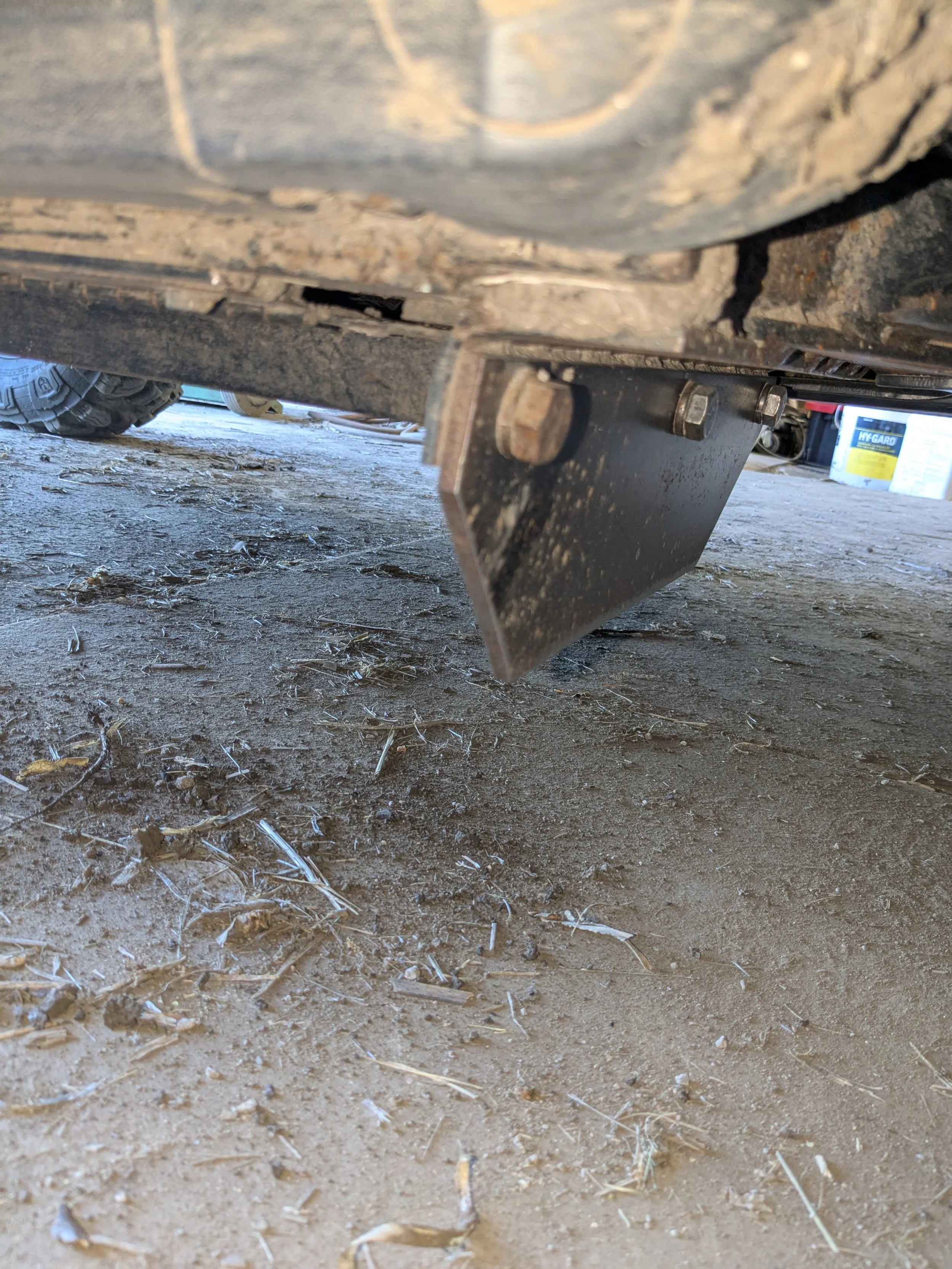 Close-up of the underside of a vehicle showing a black metal hitch receiver attached to the frame, with bolts securing it. The ground beneath is dirt with small twigs and debris.