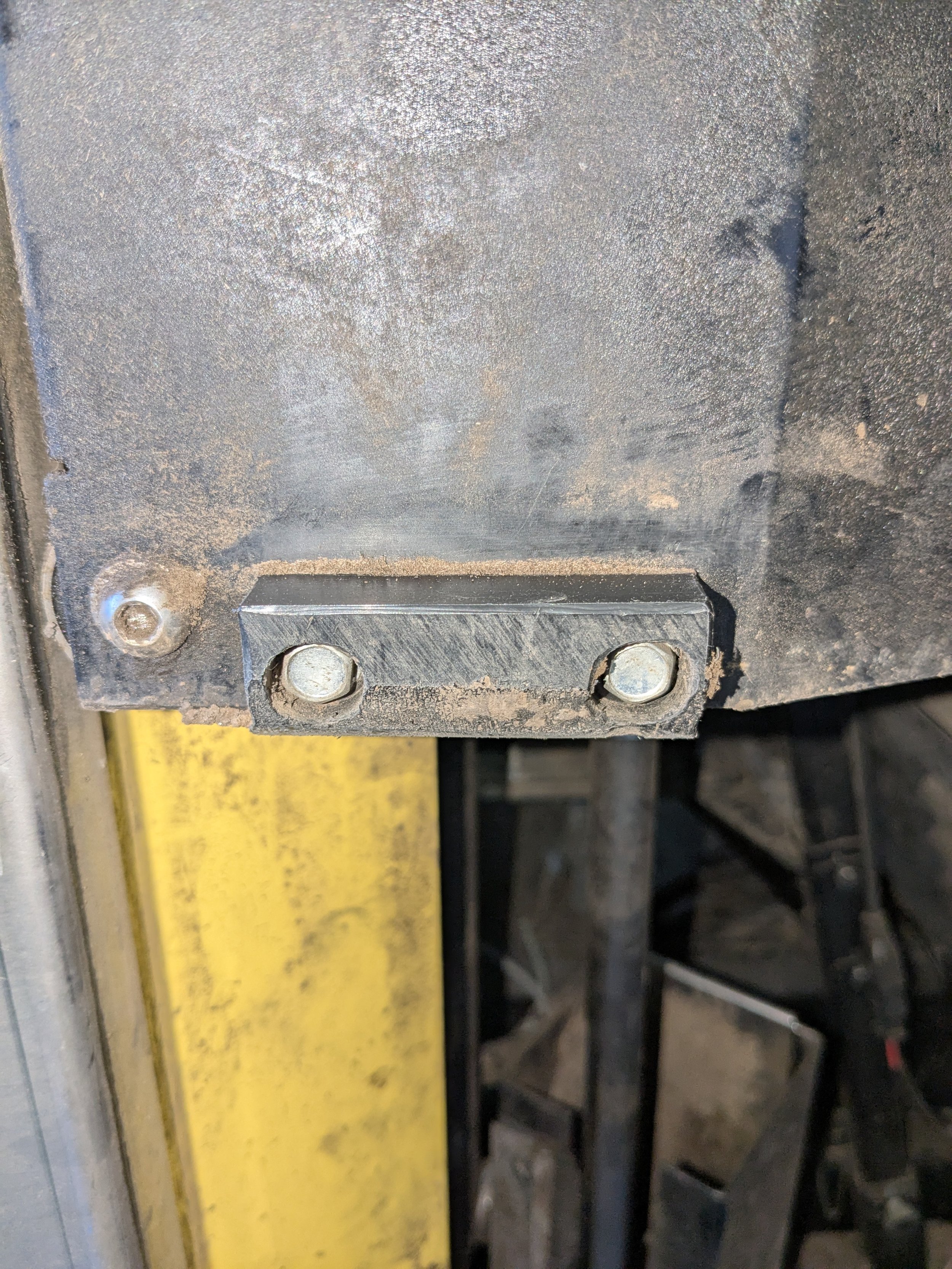 Close-up of a metal component attached with two bolts, with visible dust and dirt, against a yellow vertical structure and some black industrial equipment in the background.