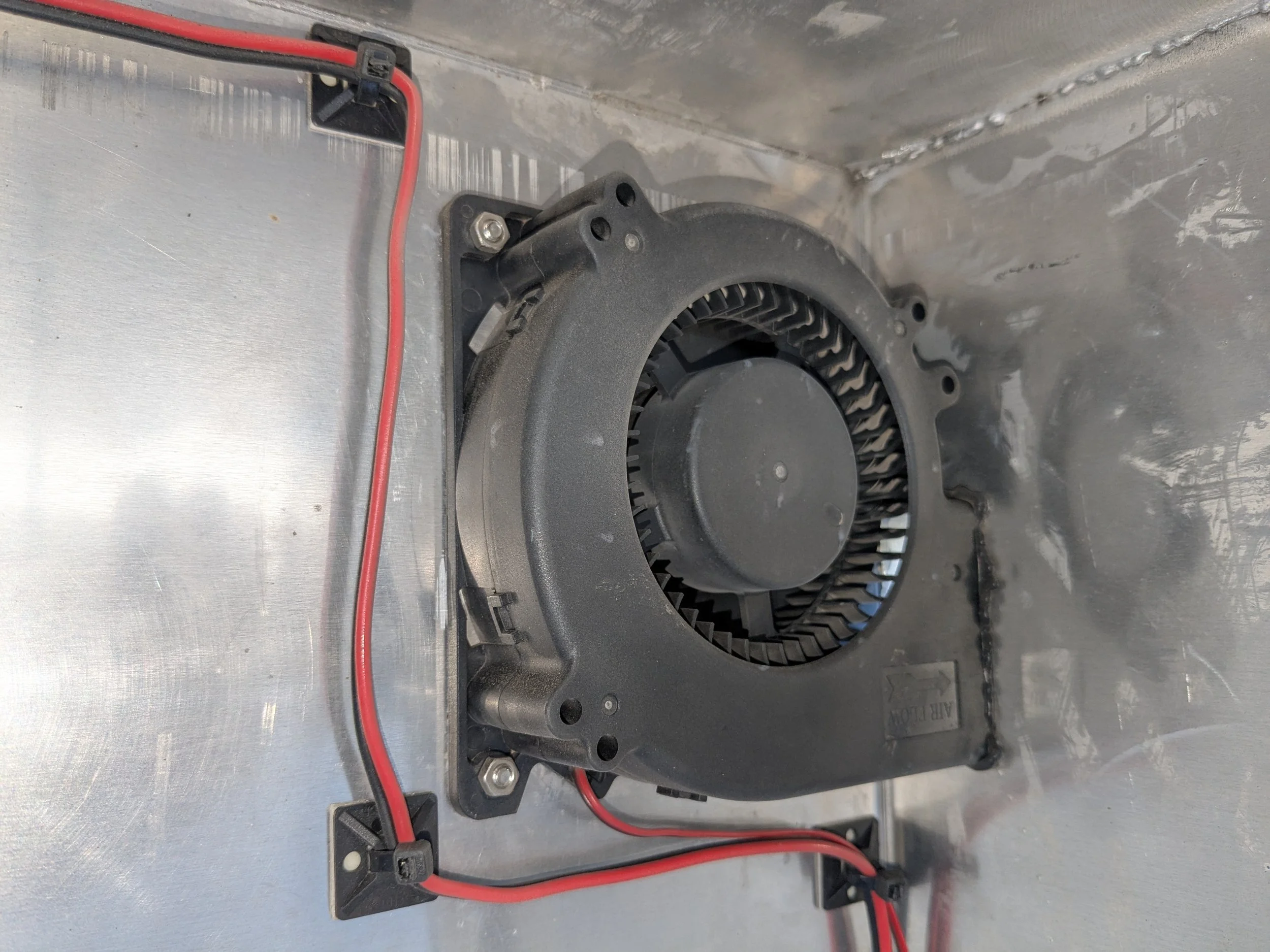 A black cooling fan or blower mounted inside a metallic enclosure with red and black electrical wires connected to it.