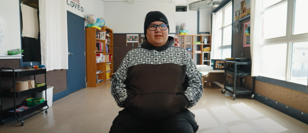 A young person sitting in a classroom with shelves, windows, and classroom supplies in the background.