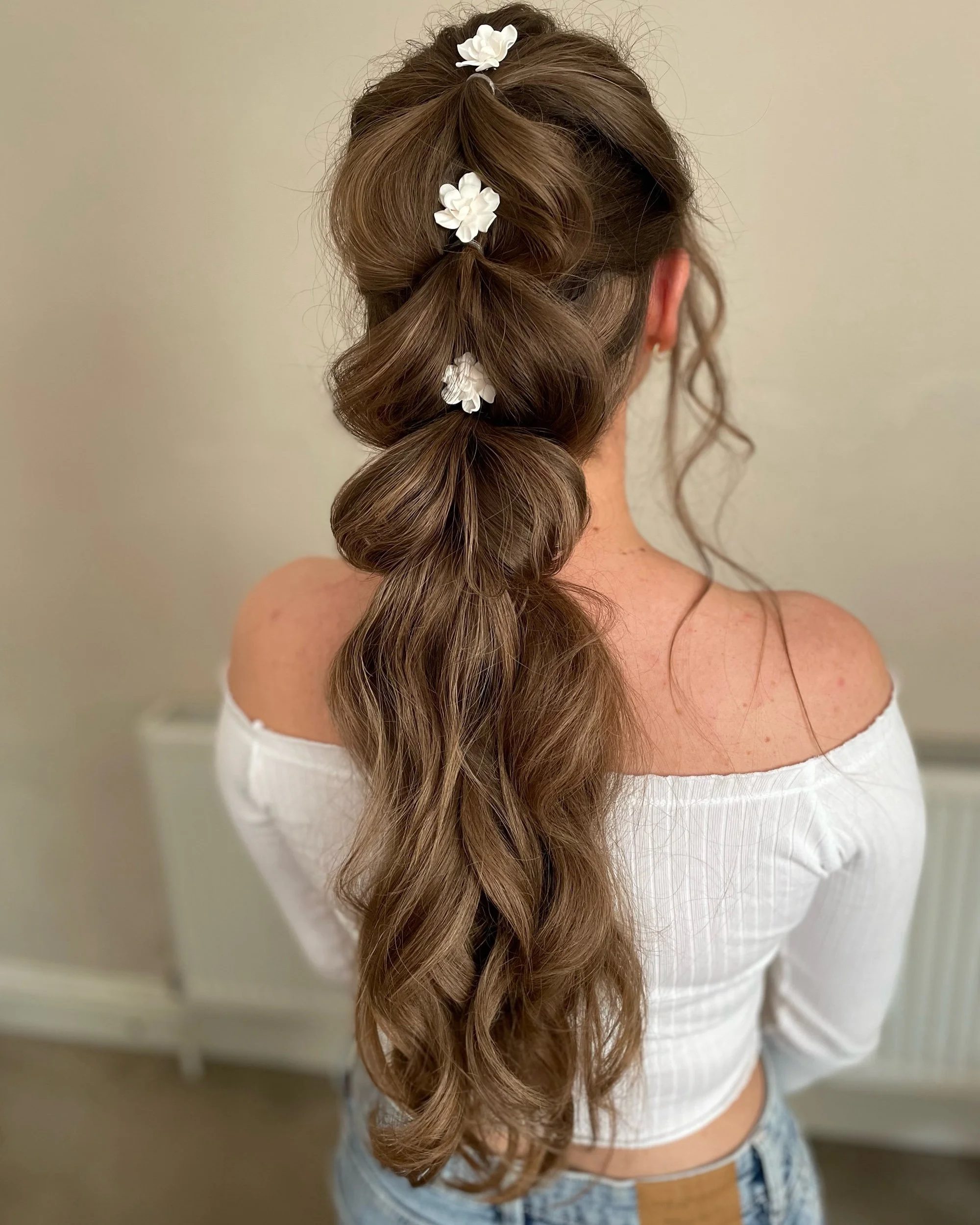 Back view of a woman with long, wavy brown hair styled in a half-up, half-down fashion with decorated white flowers.