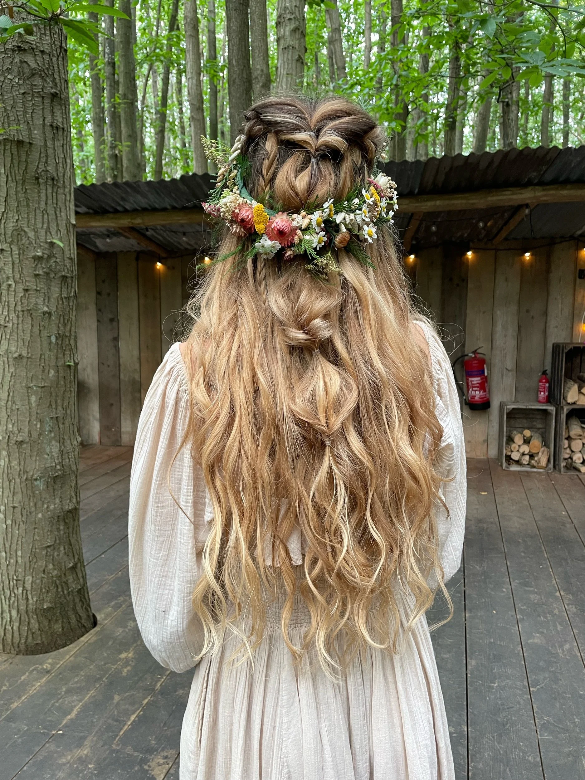 A woman with long wavy blonde hair wearing a floral crown made of colorful flowers and greenery, dressed in a light-colored peasant dress, standing outdoors near trees and a rustic wooden shed.