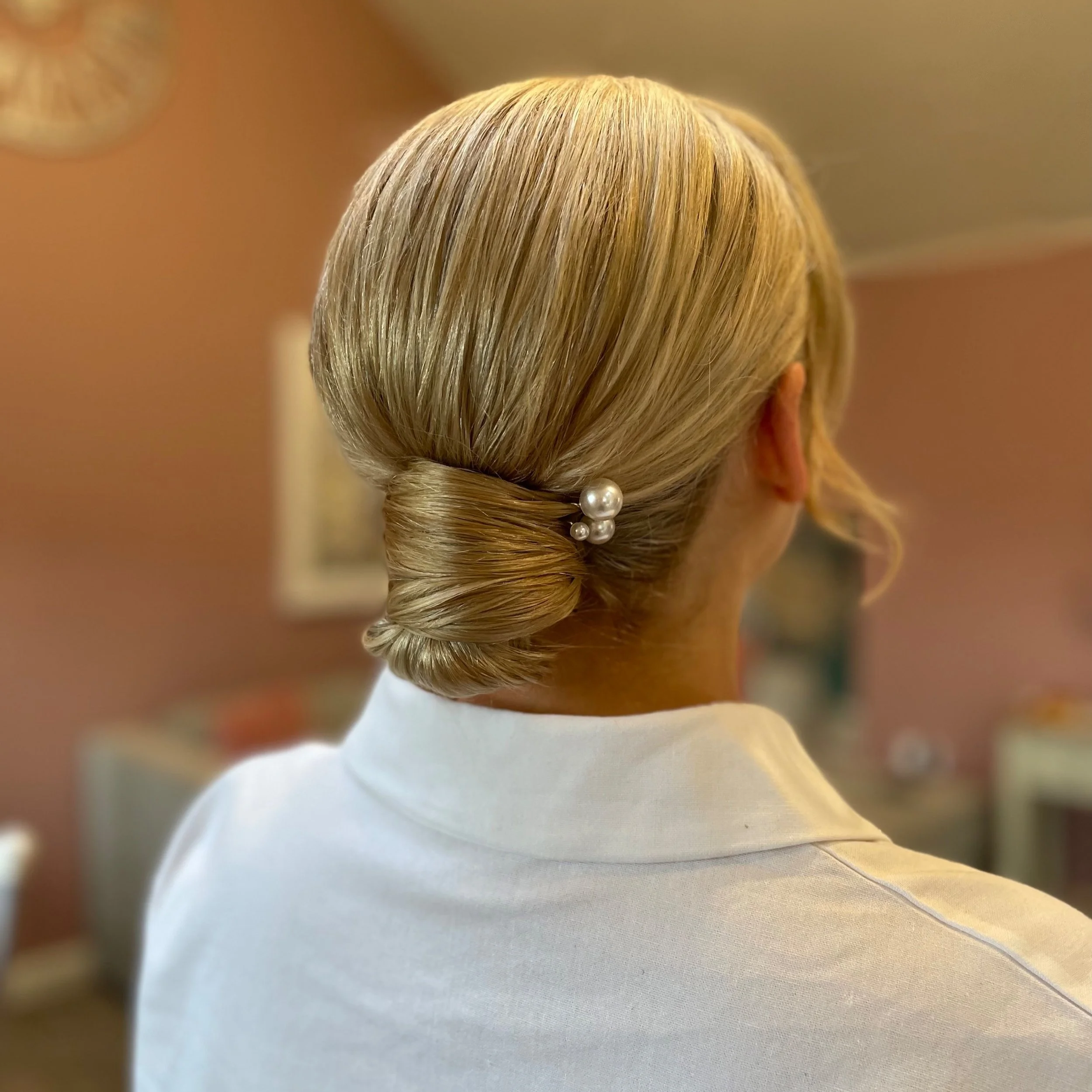 Back of a woman's head with blonde hair styled in a low bun adorned with pearl hairpins, wearing a white collared shirt, in an indoor setting.