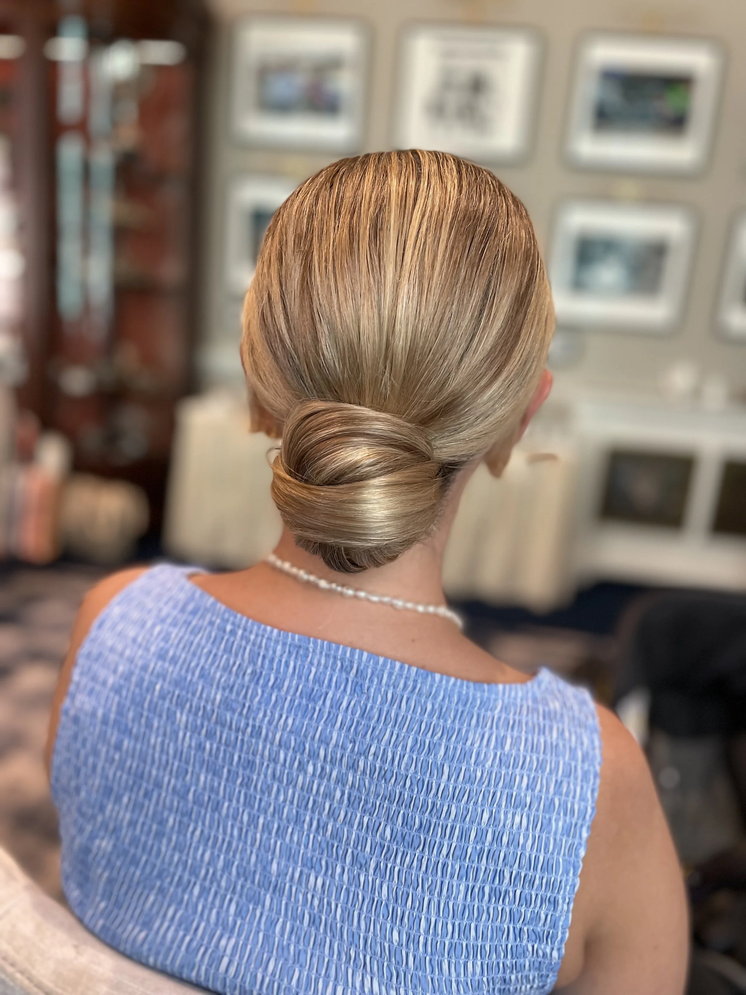 Back of a woman with blonde hair styled in an elegant low bun, wearing a blue textured sleeveless dress and a pearl necklace, in a room with framed pictures on the wall.