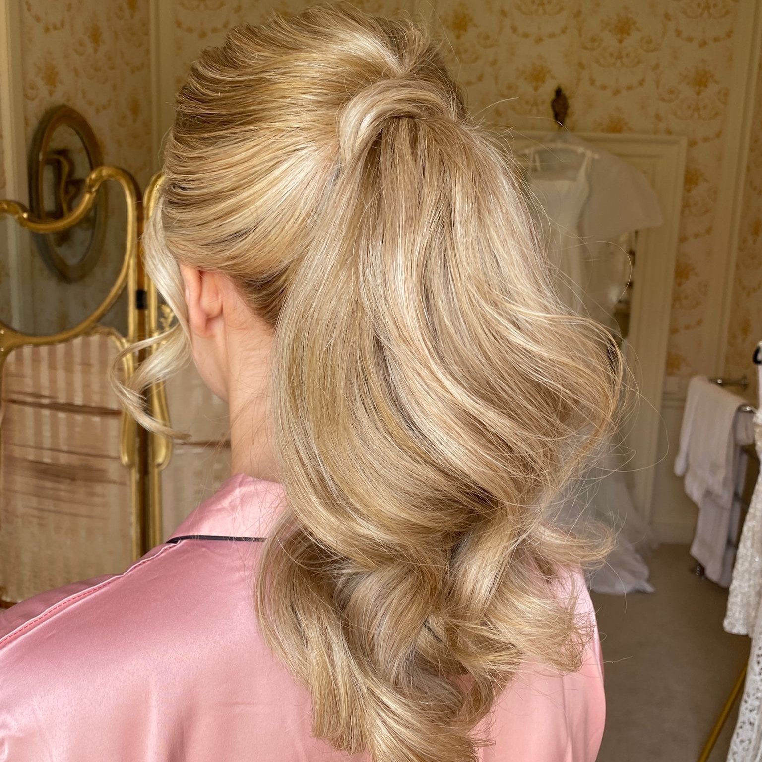 Back view of a woman with styled blonde hair in loose curls, wearing a pink satin robe, in a vintage bedroom setting.