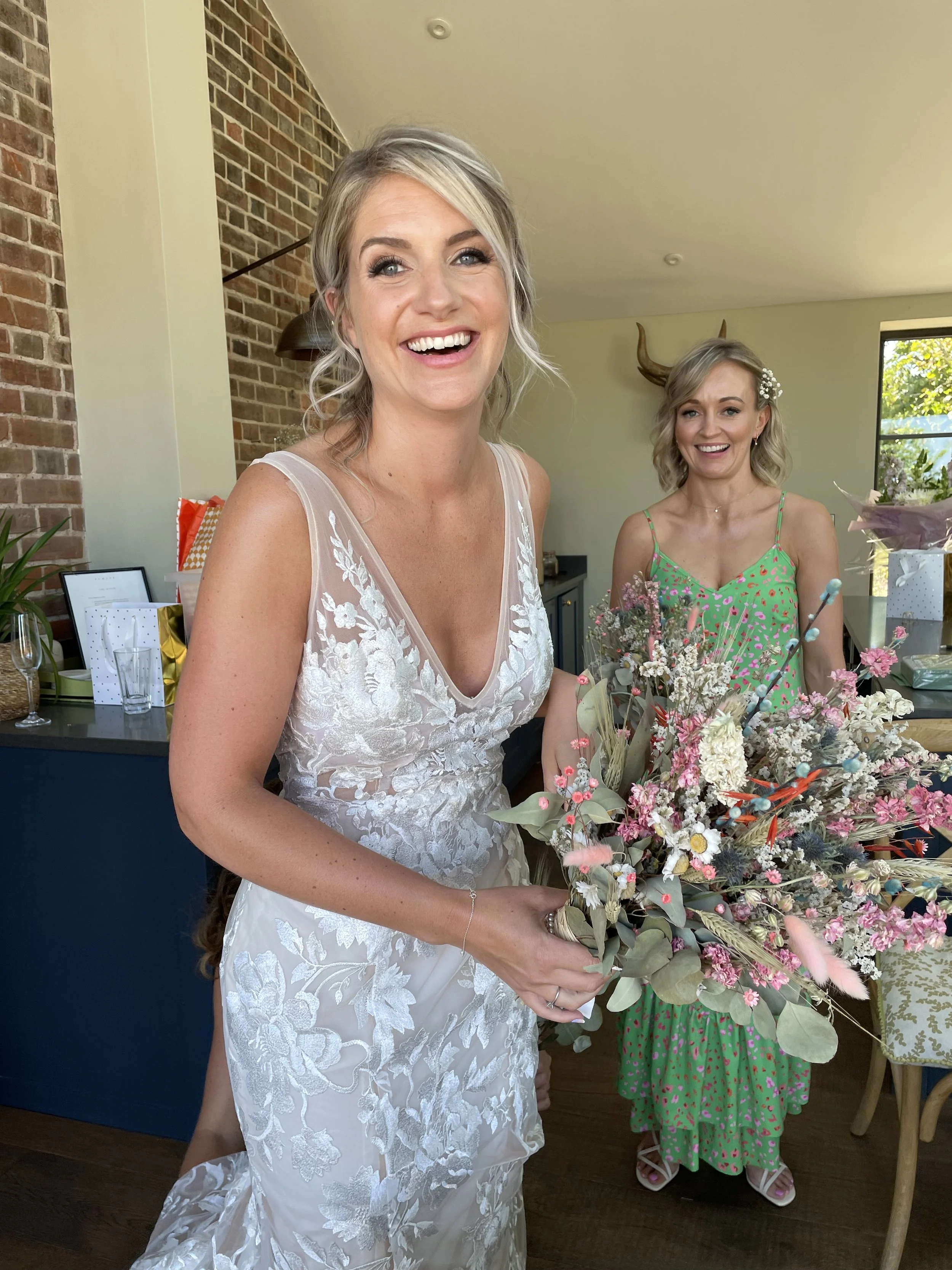 Two women in a room, one in a white wedding dress holding a bouquet of flowers, smiling, and another in a green dress with pink patterns standing nearby, with a vintage-style decor including deer antlers on the wall.