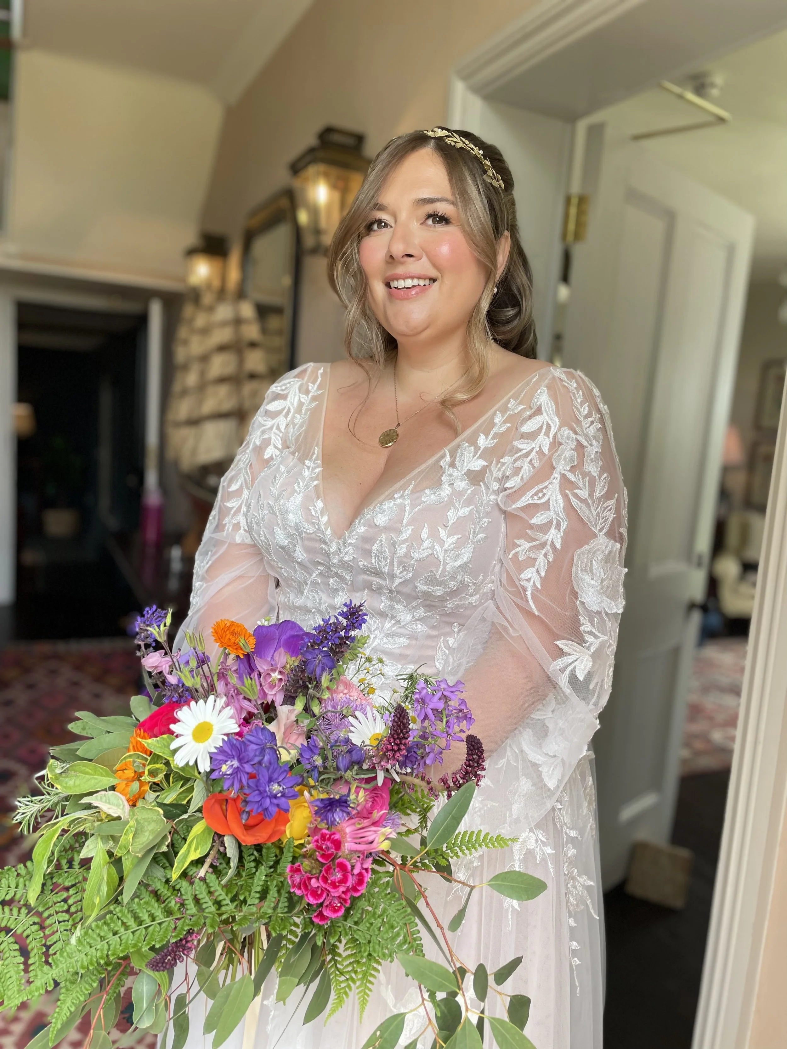 A woman in a wedding dress holding a colorful bouquet of flowers, smiling inside a room.