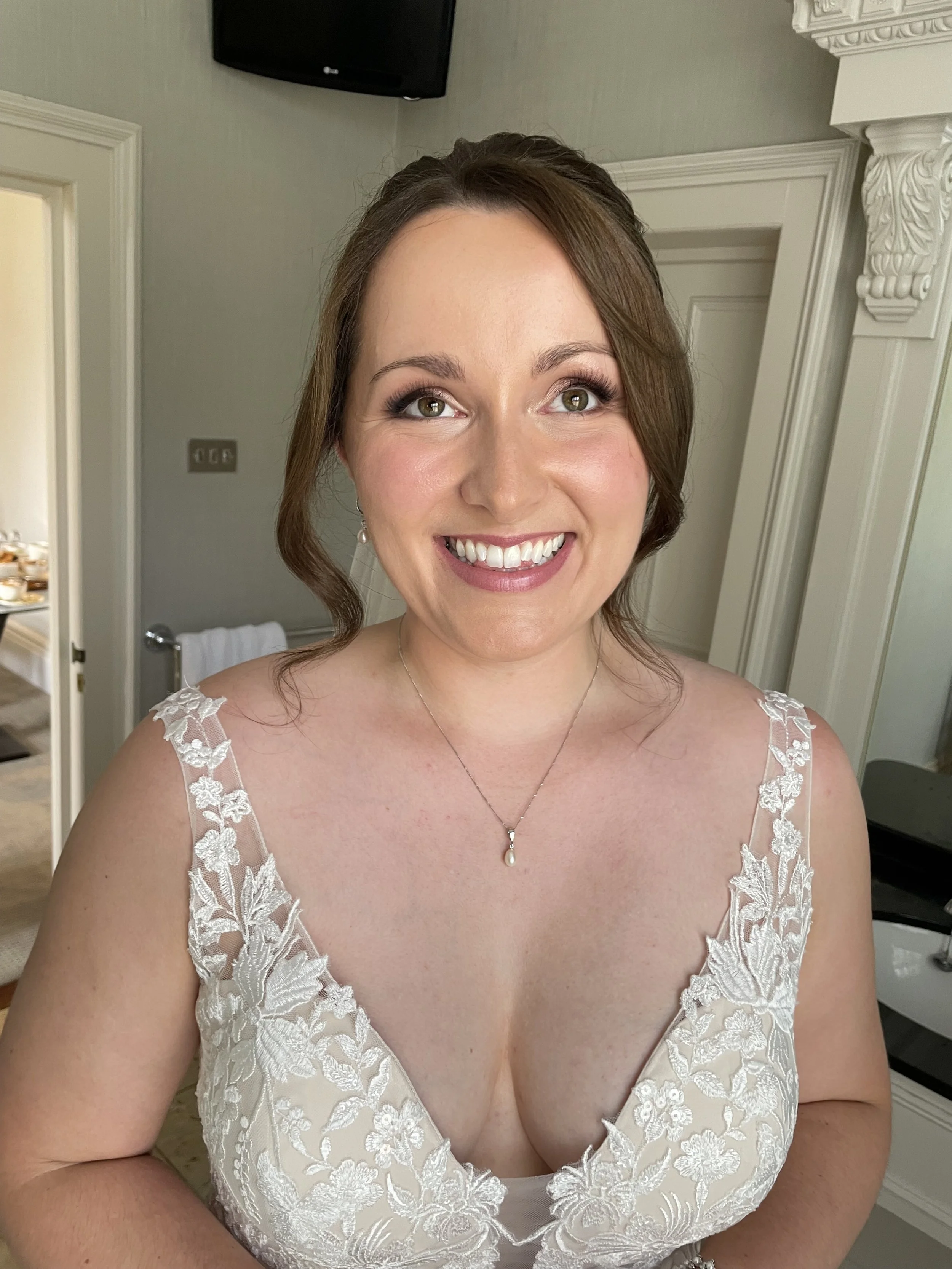 A woman with brown hair styled in loose curls, wearing a white lace dress, a pearl pendant necklace, and earrings, smiling in a room with beige walls, a door, and a TV mounted on the wall.
