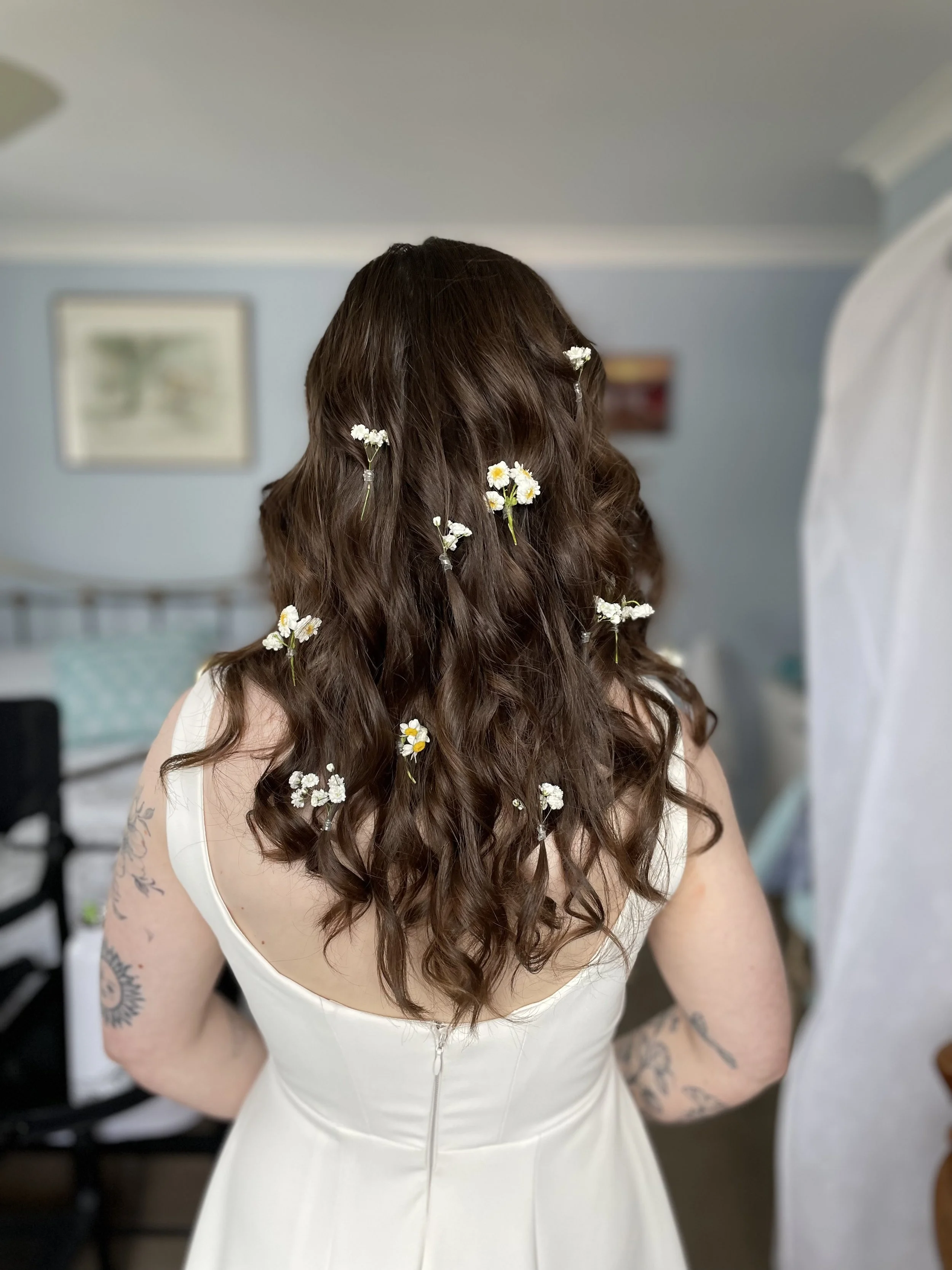 Woman with curly brown hair adorned with small white flowers, wearing a sleeveless white dress, facing away in a bedroom.