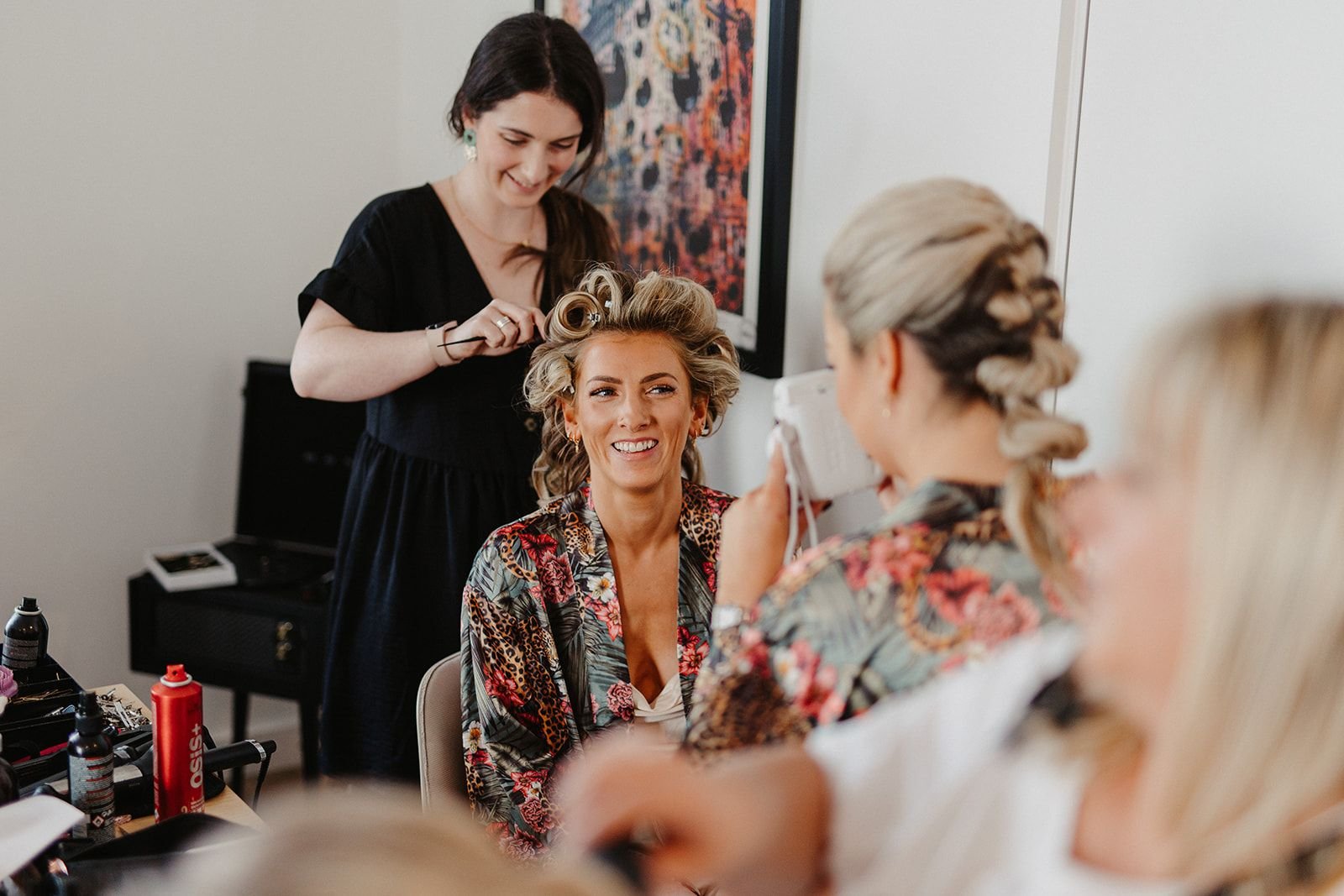 A woman with styled hair seated in a chair, smiling while a hairstylist, standing, curls her hair in rollers. Another woman, partially visible, takes a photo or videos with a camera. Several hair styling products are on a table in the foreground.