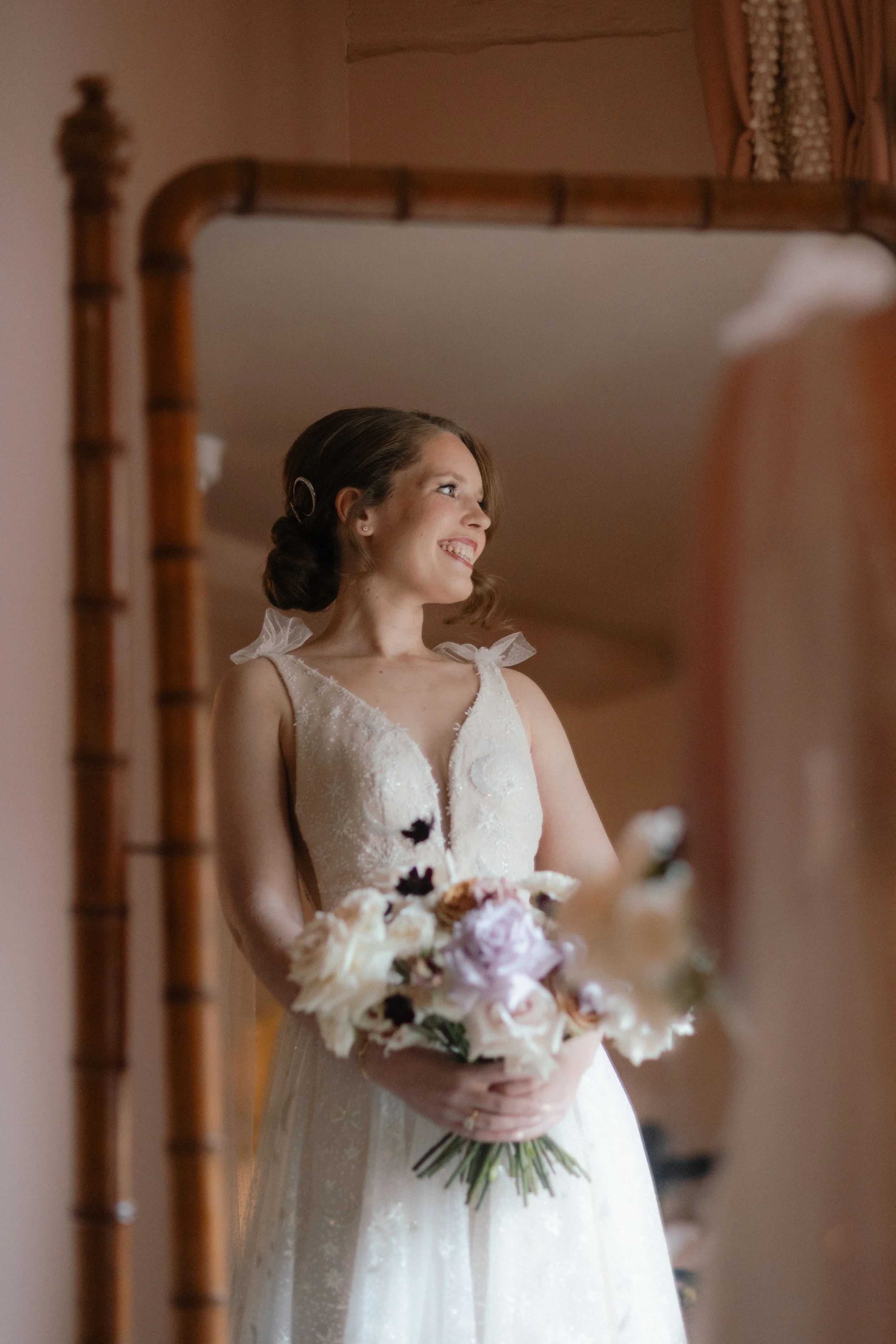 A bride in a wedding dress looking at herself in a mirror, holding a bouquet of flowers, smiling.
