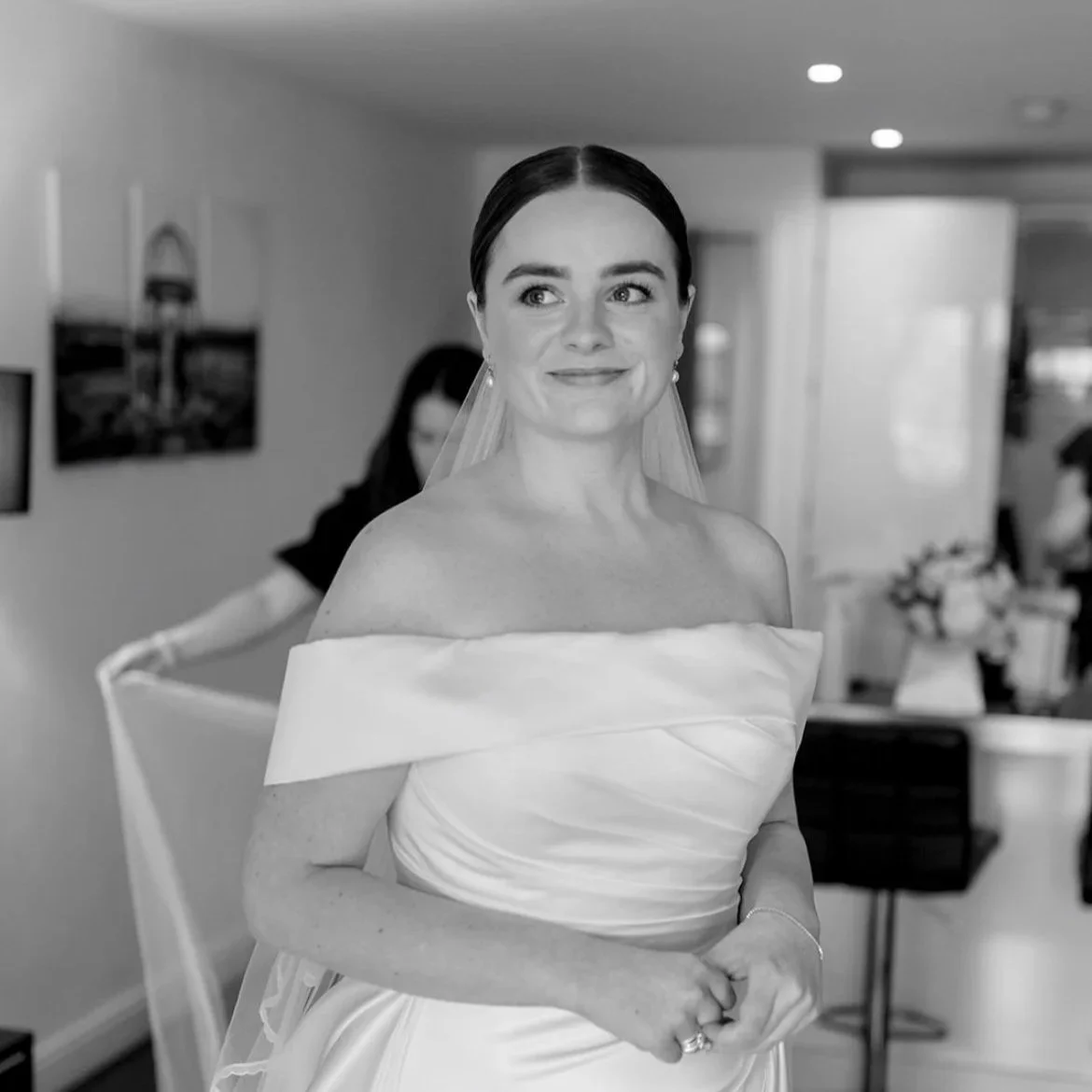 A bride in a strapless wedding gown with an off-the-shoulder neckline, smiling softly while standing indoors, with dark hair styled in an updo and a veil.