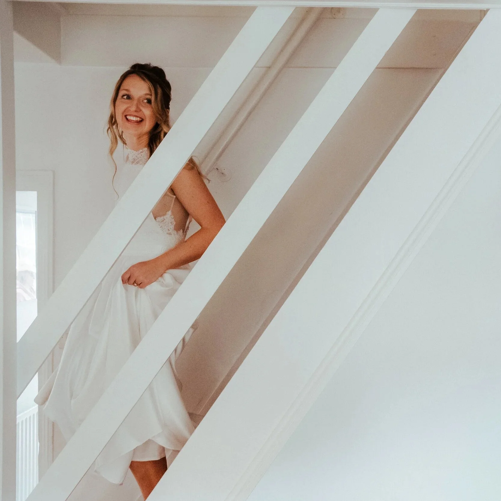 A woman in a wedding dress smiling and adjusting her dress while standing on a staircase.