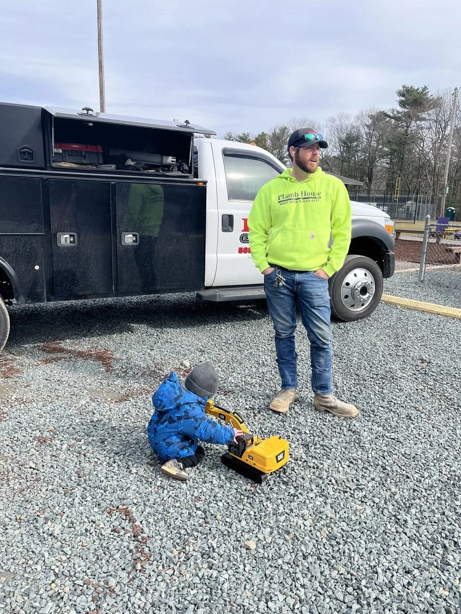 A man and a young child standing outdoors on gravel, with the child playing with a yellow toy vehicle. The man is the owner of H&M Excavation. He wears a bright green hoodie, jeans, and boots, and the child is dressed in a blue jacket and gray hat.
