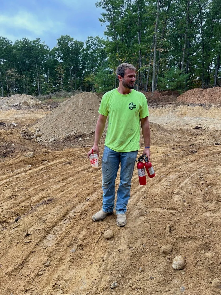 An excavation worker is wearing a bright green t-shirt and jeans standing on dirt job-site holding a red tool in one hand and a drink cup in the other. There is a background of freshly excavated dirt piles and green trees.