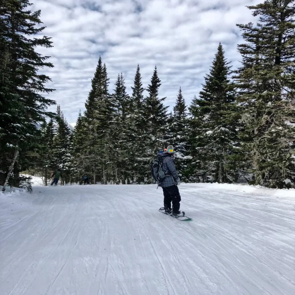 The co-founder of H&M Excavation is shown doing his out-of-work hobbies, snowboarding down on a snowy New England ski mountain surrounded by tall evergreen trees under a cloudy sky.