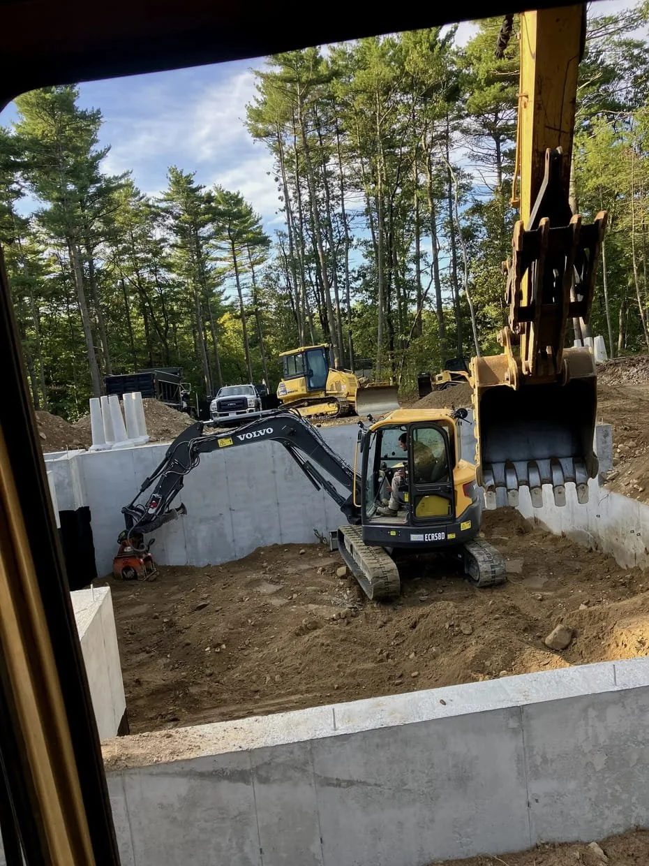 Residential construction site with a small excavator digging a new home foundation, surrounded by trees and construction equipment.